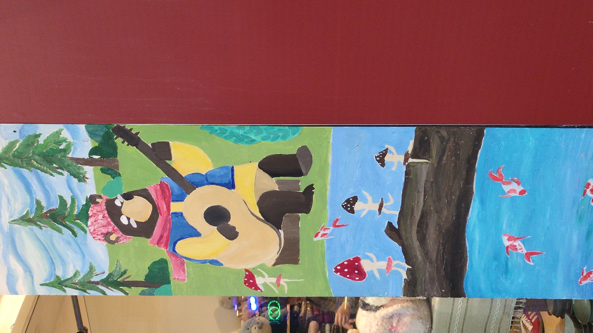 A rectangular painting depicts a stylized natural scene. On the left is a blue-and-white waterfall and green pine trees. The central portion contains a bear-like figure with a pink hat and scarf, wearing a blue jacket with a yellow shirt, seated and playing a guitar against a green background. The right side features a blue field with red and white koi fish swimming near a brown tree trunk, along with white mushrooms with red and brown spotted caps. A dark red or burgundy-colored strip appears above the painting. The painting appears to be an artwork, possibly a mural or a panel. The bottom of the image shows elements of the surroundings of the room, including a blurry display of stuffed animals and miscellaneous items. The location, Da Lat, Vietnam, is not directly evident from the visual content alone.