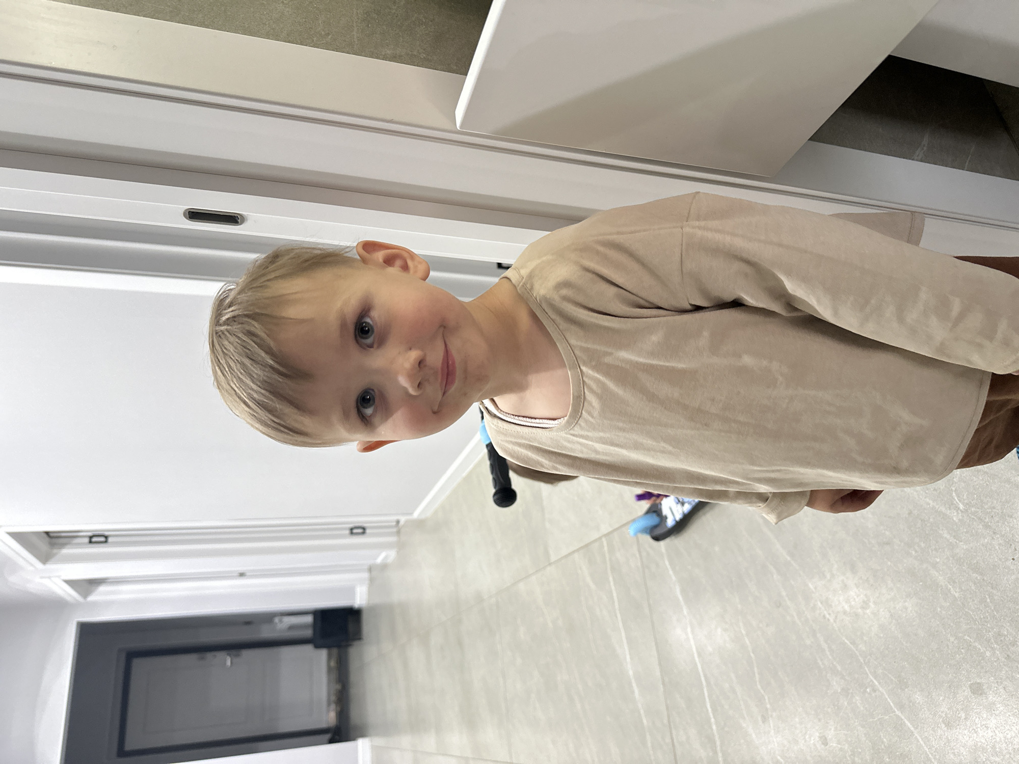 A young boy with blond hair and blue eyes stands in a hallway. He is wearing a beige long-sleeved shirt and brown pants. A scooter is visible behind him. The hallway has tiled floors and white walls with doors. The image appears to be taken indoors, possibly in a residential building, in the city of Estachi, Russia.