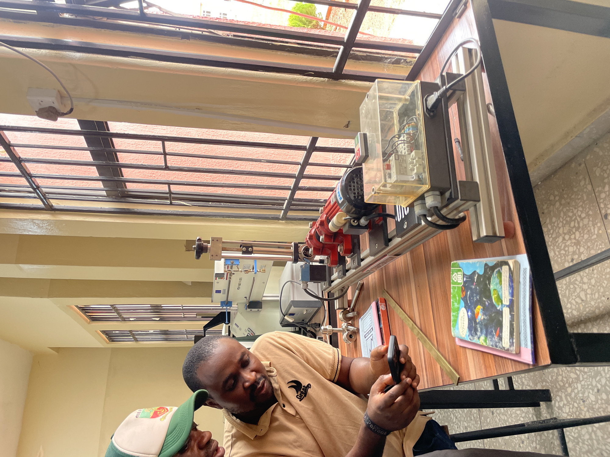 The image shows two men looking at a smartphone on a table indoors. The man in the foreground wears a beige polo shirt. A man in a green and white baseball cap is partially visible next to him. On the table are books and a red motor with a clear plastic electrical housing, part of an apparatus of some kind. The location could be Afaka, Nigeria, based on the prompt. Through the windows, a building with a red tile roof and metal security bars is visible. The interior walls are painted beige.
