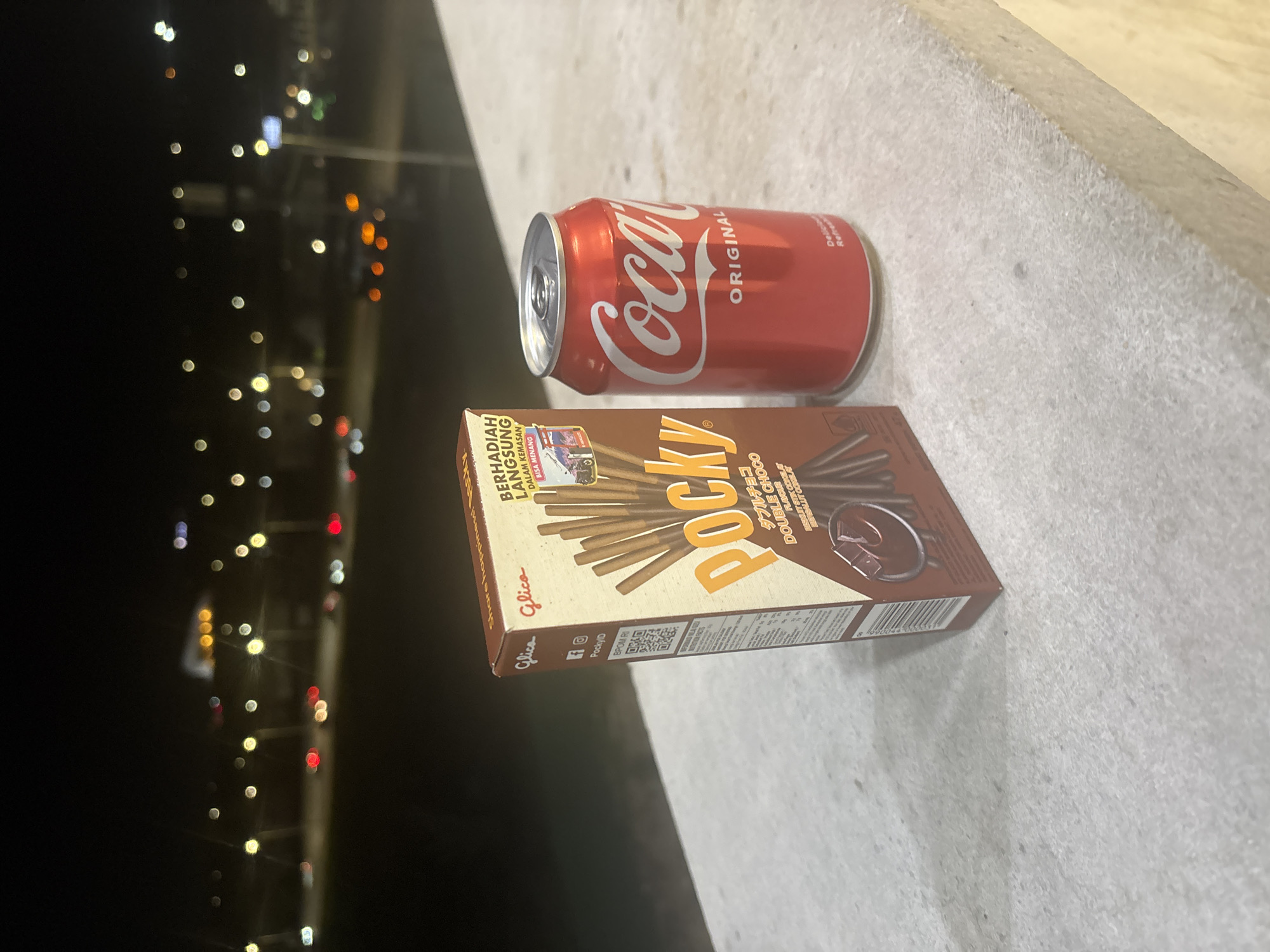 The image shows a can of Coca-Cola and a box of Pocky chocolate sticks resting on a gray, textured surface, likely a ledge or wall. In the blurred background, there are vertically aligned bokeh lights, suggesting a city scene at night. Based on visual cues like packaging and the style of the cityscape in the background, it is most likely not in Yerevan, Armenia.
