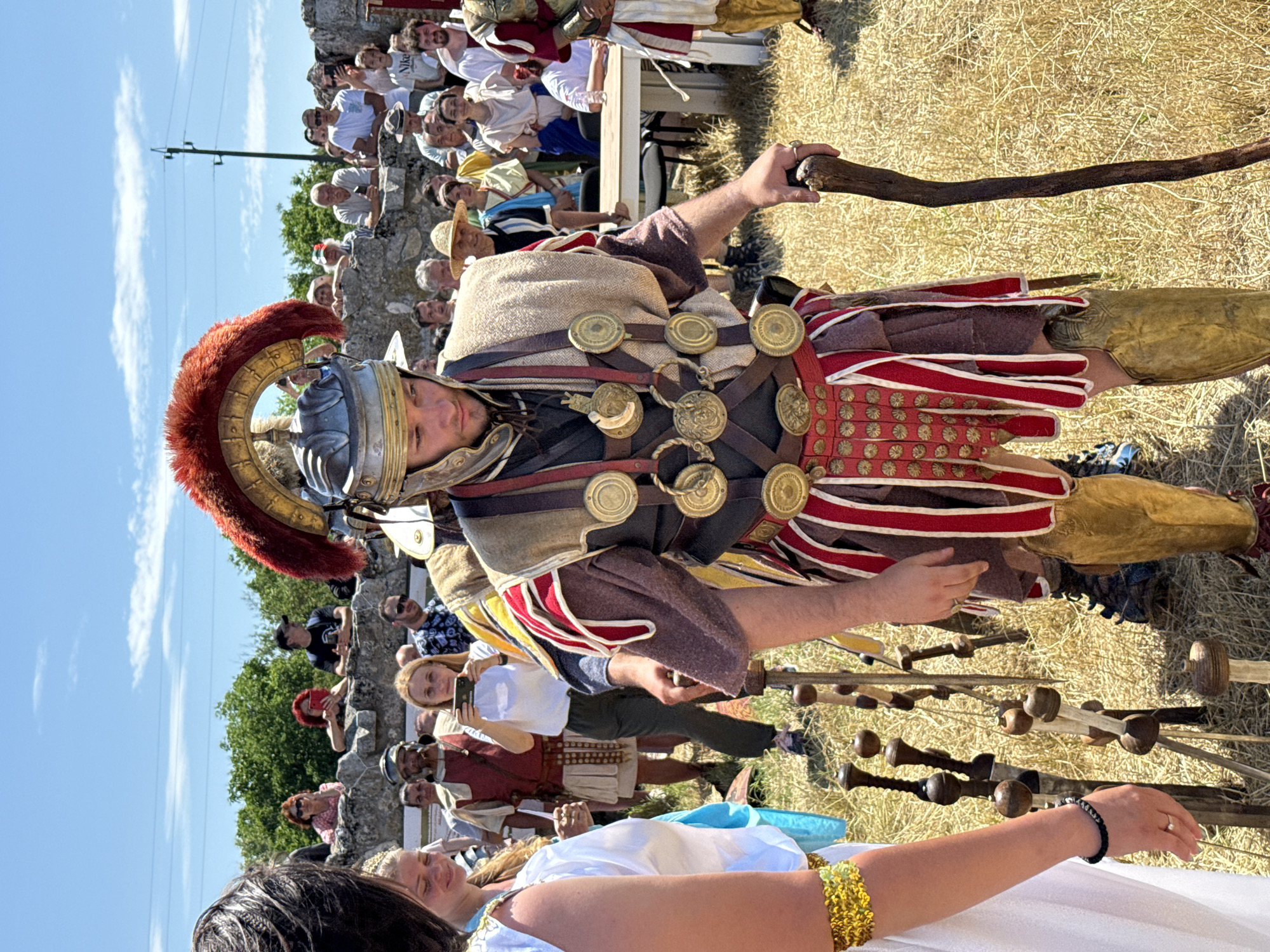 This image, captured in Gigen, Bulgaria, presents an outdoor historical reenactment or festival under a clear blue sky with scattered white clouds. The primary subject, a male reenactor in Roman military attire, occupies the center-right foreground. He wears a metal helmet featuring a prominent red horsehair crest, segmented armor adorned with multiple circular brass medallions on leather straps over a tunic, a reddish-brown tunic, red and white striped pteruges (skirt flaps), and brass greaves on his shins. He holds a dark, gnarled wooden staff, resting its base on the dry, golden-brown ground, and his gaze is directed downwards.
To the immediate left, a female figure is partially visible, wearing a white, ancient-style garment. Her left arm is adorned with a golden beaded cuff, and her left wrist displays a black beaded bracelet, with a ring on her finger.
The mid-ground and background are populated by numerous spectators. Many observers are positioned atop and behind a low, rough-hewn stone wall, which appears to be ancient ruins, covered by green trees and bushes in the distance. Other attendees are visible standing on the ground, some behind a white rectangular table with black chairs on the right. The crowd consists of individuals in both modern clothing (e.g., a man with a black "Nike" cap, women holding smartphones) and some in period-appropriate costumes (e.g., a man in a red tunic and white kilt-like garment to the right of the table, another similar figure behind the woman with the phone). Several spectators are actively photographing or video-recording the event with smartphones.
The ground surface is covered in dry, yellowish-brown grass or straw. In the immediate foreground, between the two main figures, several upright wooden staffs with distinctive spherical tops are planted in the ground. A utility pole with power lines is visible against the sky on the upper right side of the frame.