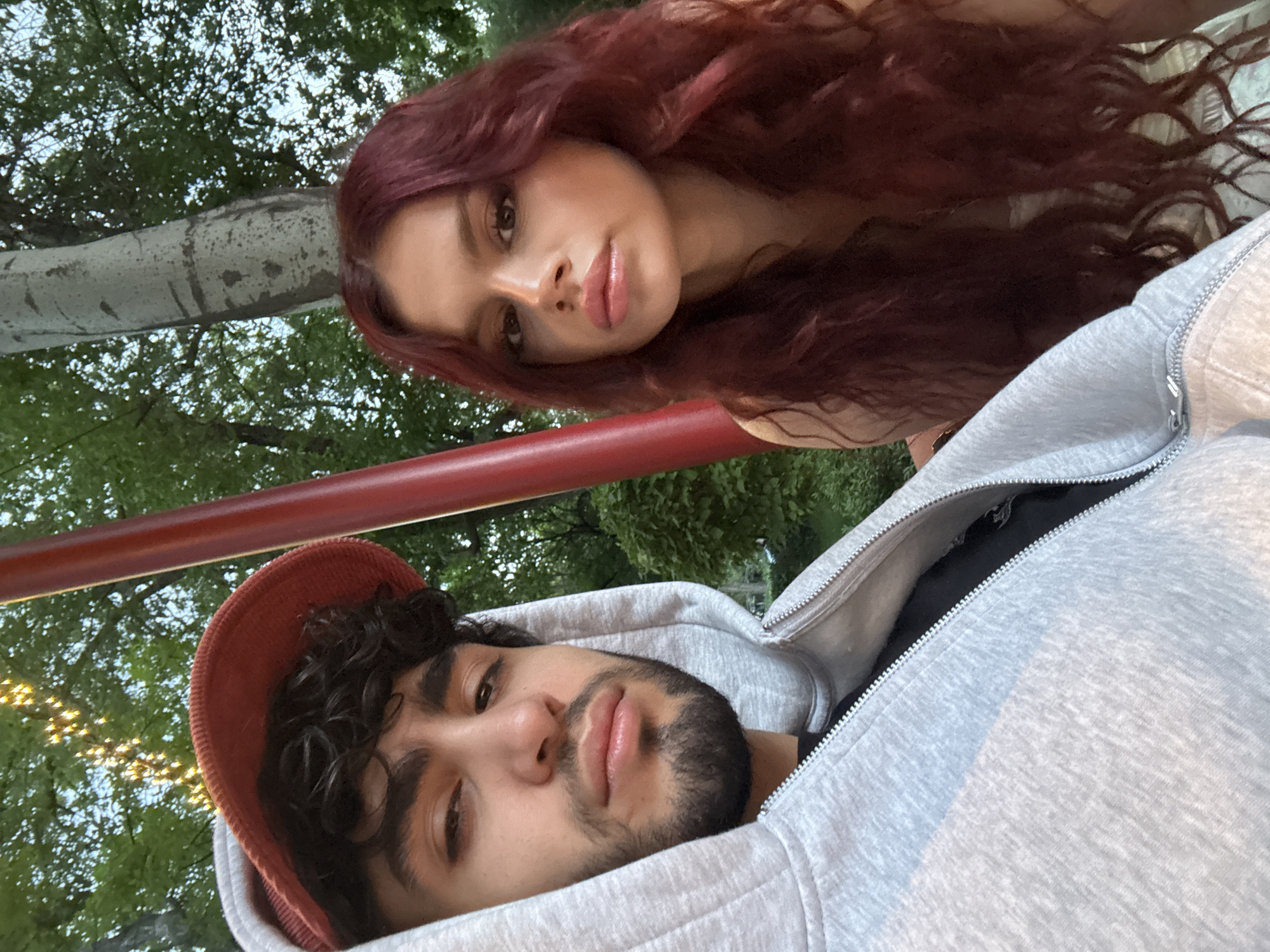 An eye-level portrait shot captures two individuals, a man and a woman, positioned at an angle, likely taken outdoors in Yerevan, Armenia. The woman is in the upper portion of the frame, lying on her side, with her head seemingly resting on her hand. She has long, wavy, reddish-brown hair and is looking directly at the camera with a neutral expression and slightly glossy lips. She appears to be wearing a light-colored top.
Below her, and slightly in front, is a man, also angled, looking towards the camera. He has dark, curly hair, a beard, and a mustache. He is wearing a grey hooded sweatshirt with a visible zipper and a rust-orange or burnt-orange colored cap. His expression is also neutral, with a slight pout to his lips.
A red metal bar runs horizontally through the mid-ground, positioned between the two subjects. In the background, lush green trees and foliage are visible under a bright sky, suggesting an outdoor park or recreational area. The lighting indicates it is likely late afternoon or early evening, with soft, natural light illuminating their faces. No text is visible in the image.