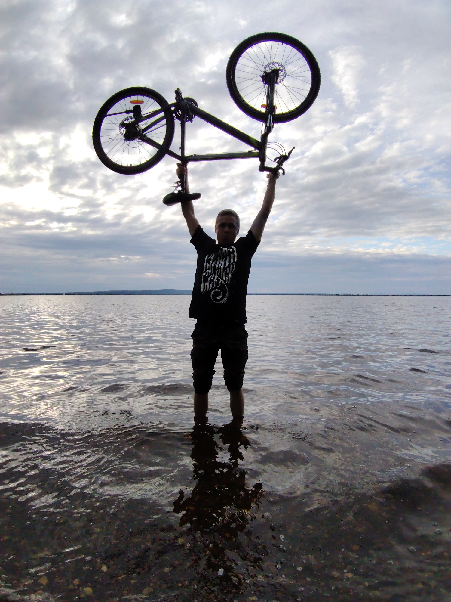 A man stands waist-deep in calm water, holding a bicycle overhead with both hands. The bicycle is silhouetted against a cloudy sky. The man is wearing a dark t-shirt and shorts. His reflection is visible in the water, as are pebbles on the lakebed. The scene appears to be set at dusk or dawn, given the soft light and cloud cover. The overall mood is one of quiet accomplishment or triumph, with the man and his bicycle presented as a unified whole against the vastness of the water and sky. The text on the man's t-shirt is indistinct but appears to be in a decorative, possibly gothic script.