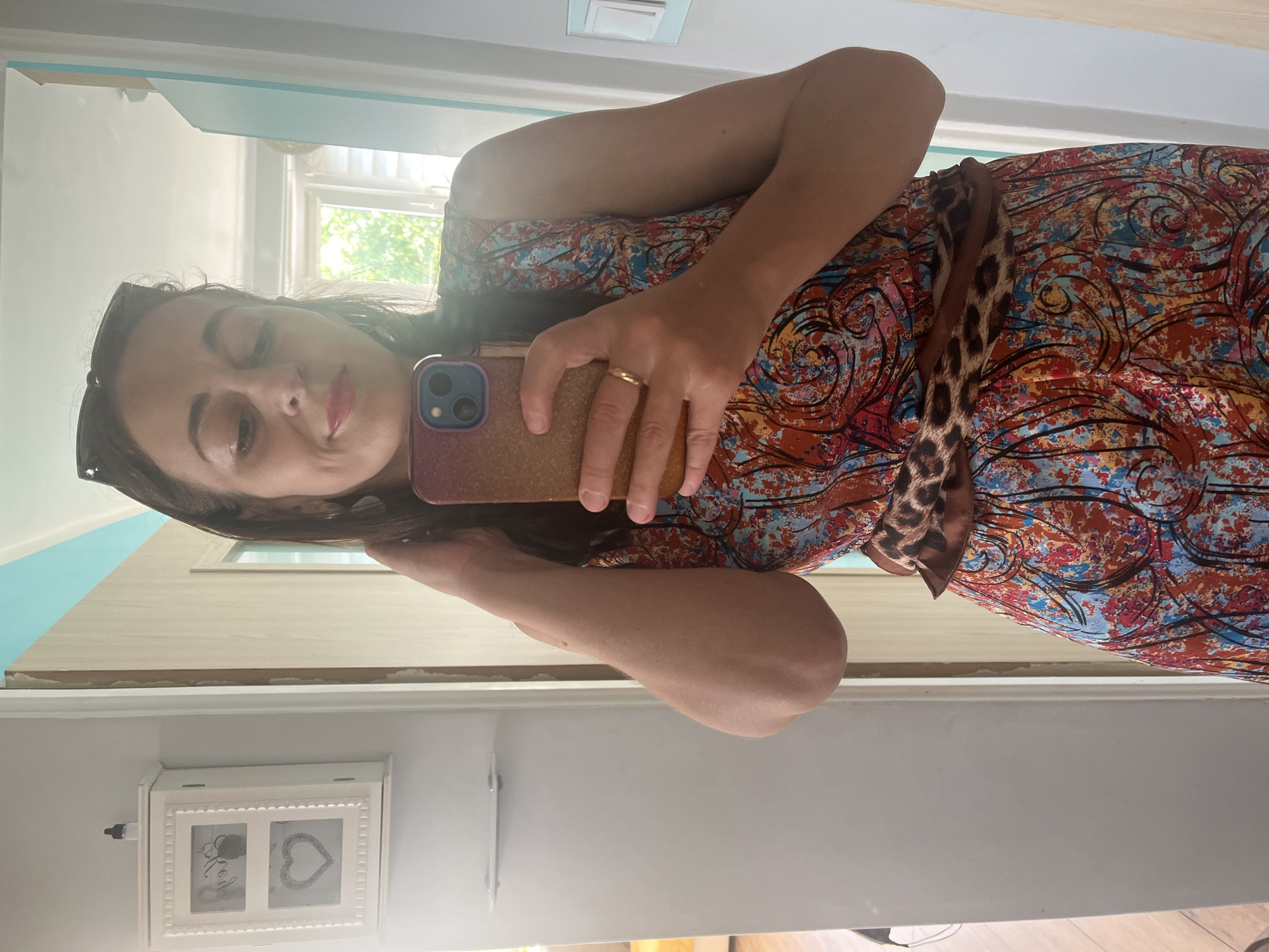 A woman is standing in front of a mirror, taking a selfie. She is wearing a colorful dress with a leopard print belt. She is holding a phone in her right hand and touching her hair with her left hand. The background is a room with a white cabinet on the left and a door on the right. There is a window in the background with greenery visible outside.