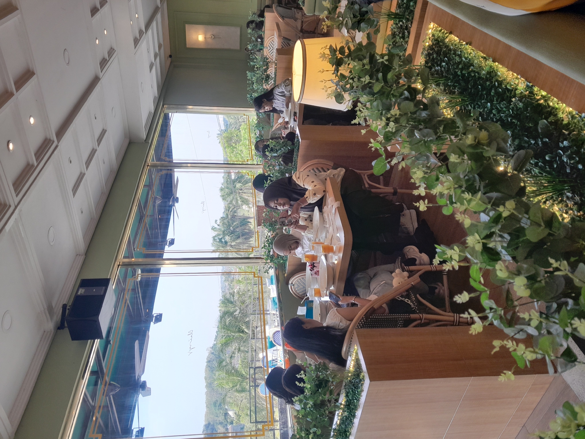 This image depicts the interior of a restaurant in Sempusari, Indonesia, with large windows offering a view of a lush, green landscape with palm trees. The restaurant's decor features light green walls, a white paneled ceiling with recessed lighting, and wooden accents. Several groups of people are seated at tables, engaged in dining and conversation. In the foreground, a group of young women is seated at a table, some looking at their phones. Another table in the mid-ground shows a woman in a headscarf eating. The tables are set with plates, bowls, and glasses of orange juice. Decorative plants with small lights are placed around the seating areas, creating a pleasant ambiance. Overhead, a black speaker is mounted on the wall. The overall scene suggests a casual dining experience in a well-lit and aesthetically pleasing environment.