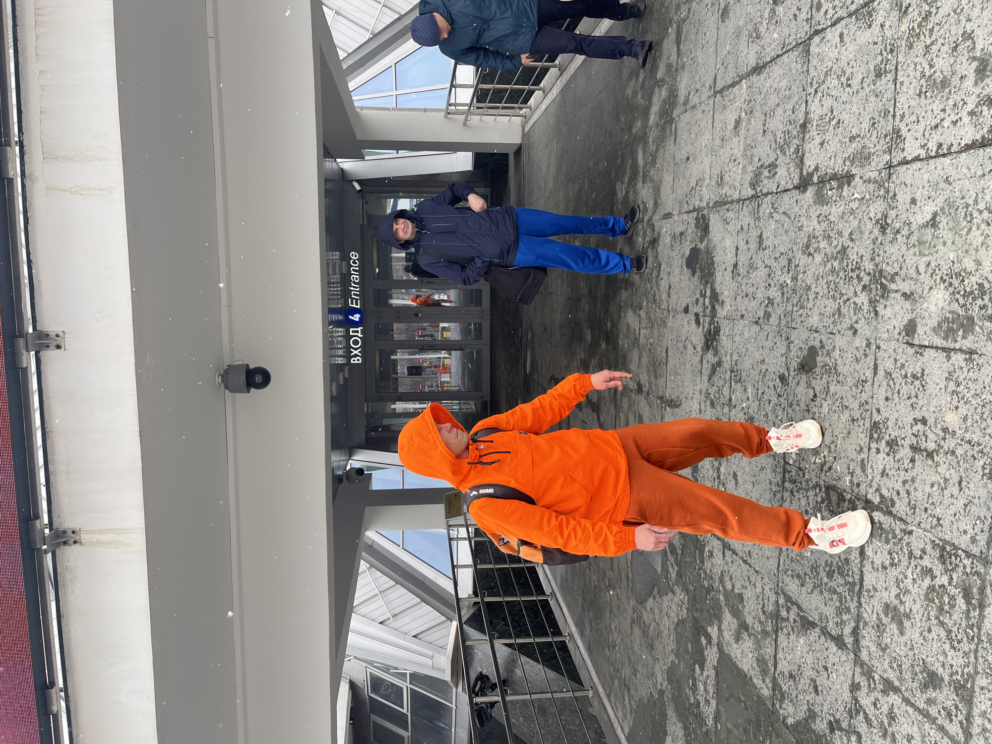 Three men are in front of an entrance with a sign written in Russian and English that reads "Entrance". The man closest to the viewer is wearing an orange hooded jacket and orange pants. The second man is wearing a blue jacket and blue pants. The third man is wearing a dark blue jacket and pants. The entrance appears to be to a building in Moscow, Russia. The ground is a grey stone. It is snowing.
