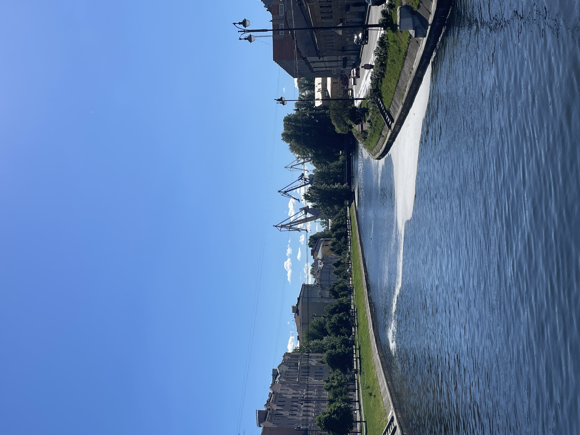 A bright, clear day with a deep blue sky illuminates a wide canal in Saint Petersburg, Russia. The canal's dark blue water is mostly calm, with ripples and a patch of foamy white material near the right bank. On the left bank, a row of multi-story buildings with grey facades and ornate architectural details lines a tree-filled embankment. A paved walkway separates the buildings from the canal. On the right bank, a street runs parallel to the canal, with several cars and a few pedestrians visible. Mature trees and green grass border the street. In the background, several tall, industrial cranes stand against the sky, suggesting a port or industrial area. The overall scene depicts an urban waterway with a mix of residential and industrial elements under a sunny sky.
