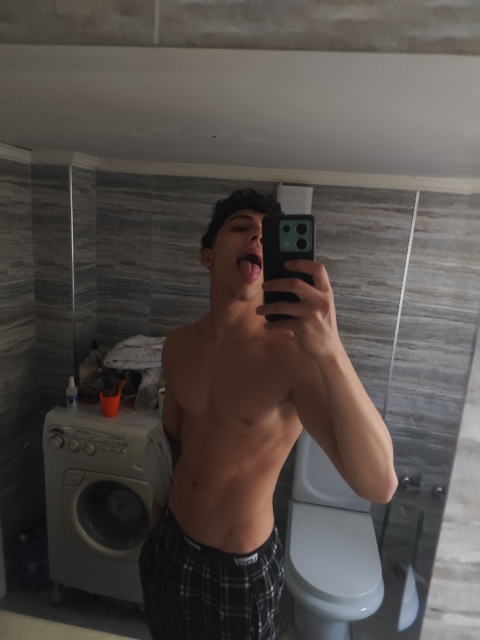 The image depicts a mirror selfie taken by a young male within a domestic bathroom setting. The specific location of As-Salihiya Municipality, Syria, cannot be verified from the visual information provided.
The subject is a young male with dark, curly hair, bare-chested, and wearing black and white plaid pajama bottoms. He is positioned centrally in the frame, holding a dark-colored smartphone with a prominent light green camera module in his right hand to capture his reflection. His tongue is extended out of his mouth.
The background reveals typical bathroom fixtures and objects. To the left of the subject, a white front-loading washing machine is visible, upon which an orange plastic cup containing multiple toothbrushes, a white spray bottle, and folded white fabric are placed. To the right of the subject, a white toilet with its lid closed is partially visible. The walls are covered in light grey tiles featuring horizontal striations, and the ceiling is plain white. The flooring is not discernible.