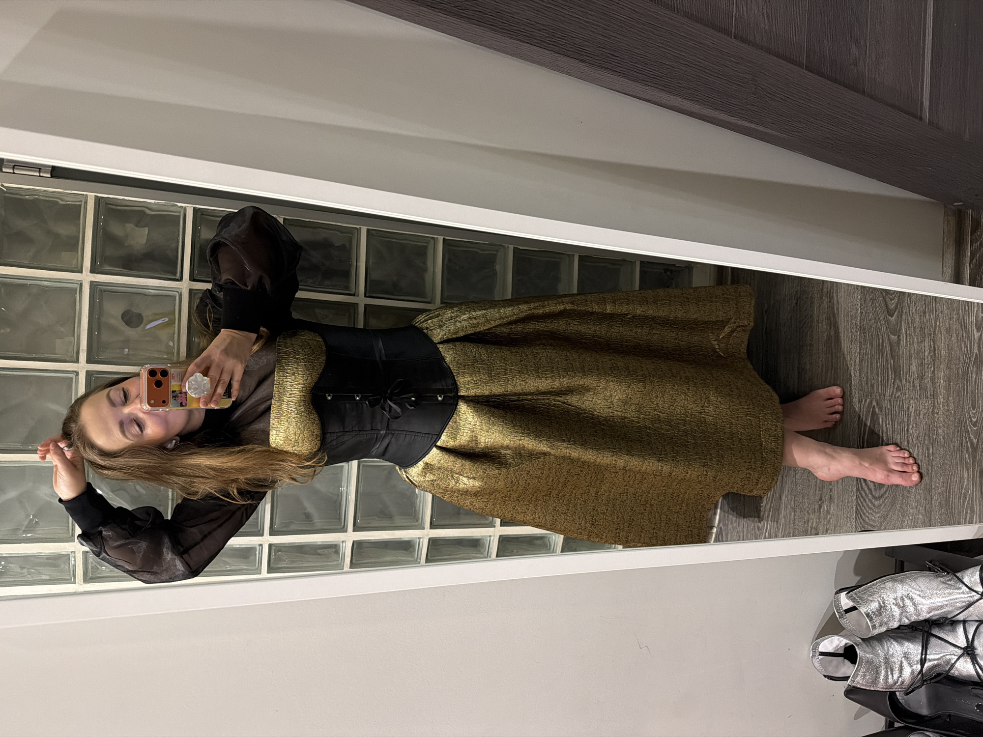 A woman is standing in front of a mirror, holding a mobile phone and taking a selfie. She is wearing a sheer black long-sleeve top, a black corset, and a gold-colored skirt that reaches below the knee. The background behind her features a wall of glass blocks. To the right, there is a dark-colored wooden wall. At the bottom right of the image, there are several pairs of silver boots stacked together. The setting appears to be indoors, likely in a residential space, in the city of Unknown, Unknown.