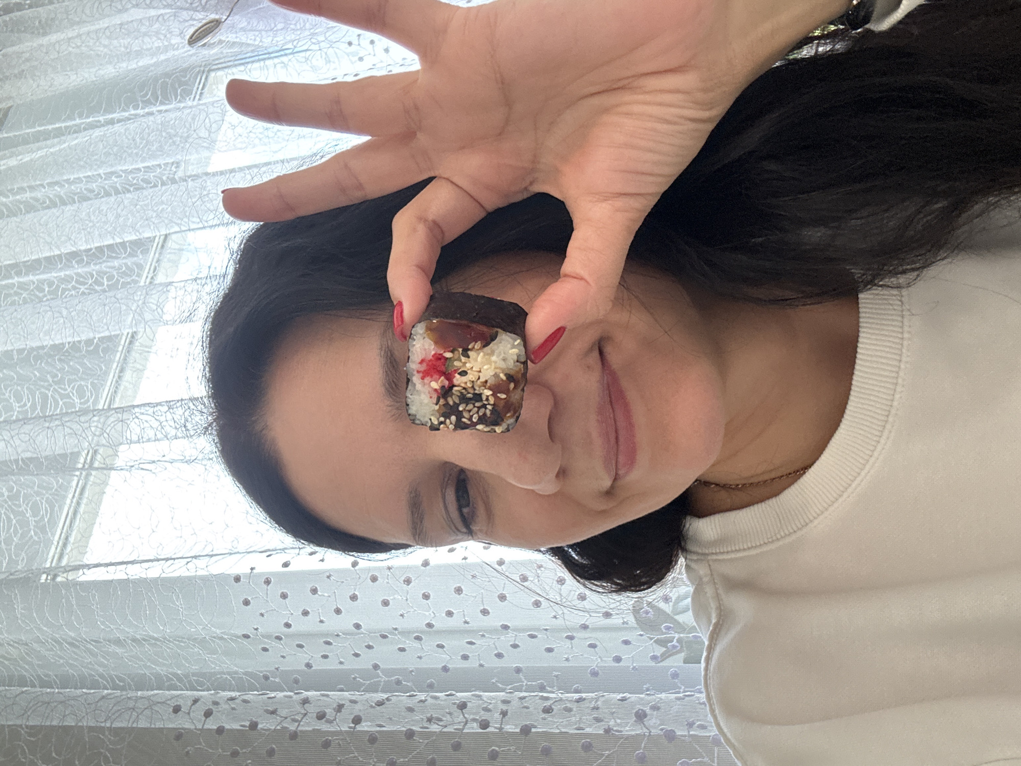 A woman is holding a piece of sushi in front of her eye. She has dark hair and is wearing a white shirt. The sushi roll appears to have rice, nori (seaweed), and some fillings. The background includes a window with patterned curtains or blinds. The image was taken from a low-angle perspective. There's no information to definitively ascertain the location is in Chaikovsky, Russia.