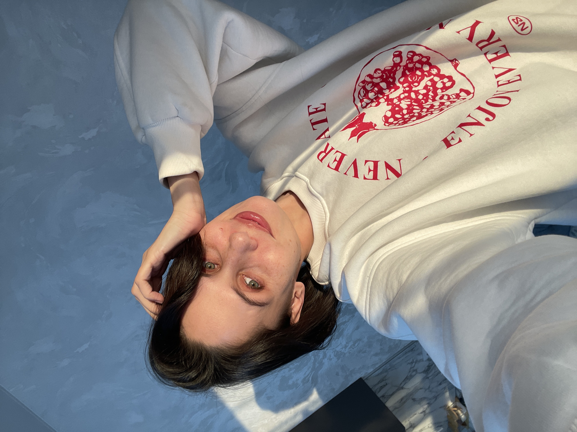 A woman with fair skin, dark hair, and green eyes is reclining on a light blue surface, possibly a wall or floor, with streaks and patterns that resemble clouds. She is wearing a white sweatshirt that features a red pomegranate design and the words "ENJOY EVERY NEVER ATE". The woman's right hand is touching her forehead, and she is looking at the camera with a slight smile. The environment includes what appears to be a section of marble-like flooring at the bottom of the image. The location is not possible to determine in Izhevsk, Russia or any city, based solely on the visual information provided.