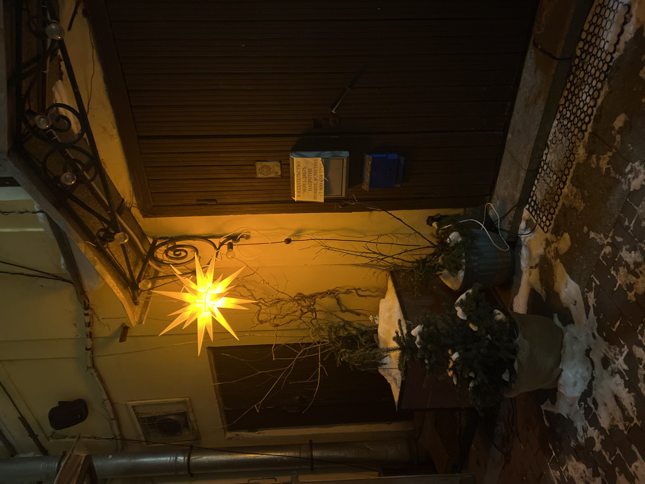 A dark, angled shot shows a building exterior at night. A bright, star-shaped light hangs from an ornate ironwork bracket. Above the light are dark brown wooden doors and an off-white wall. Potted plants with snow sit on the ground next to the doors. A black metal grate covers part of the ground. The lighting is dim except for the radiant star.