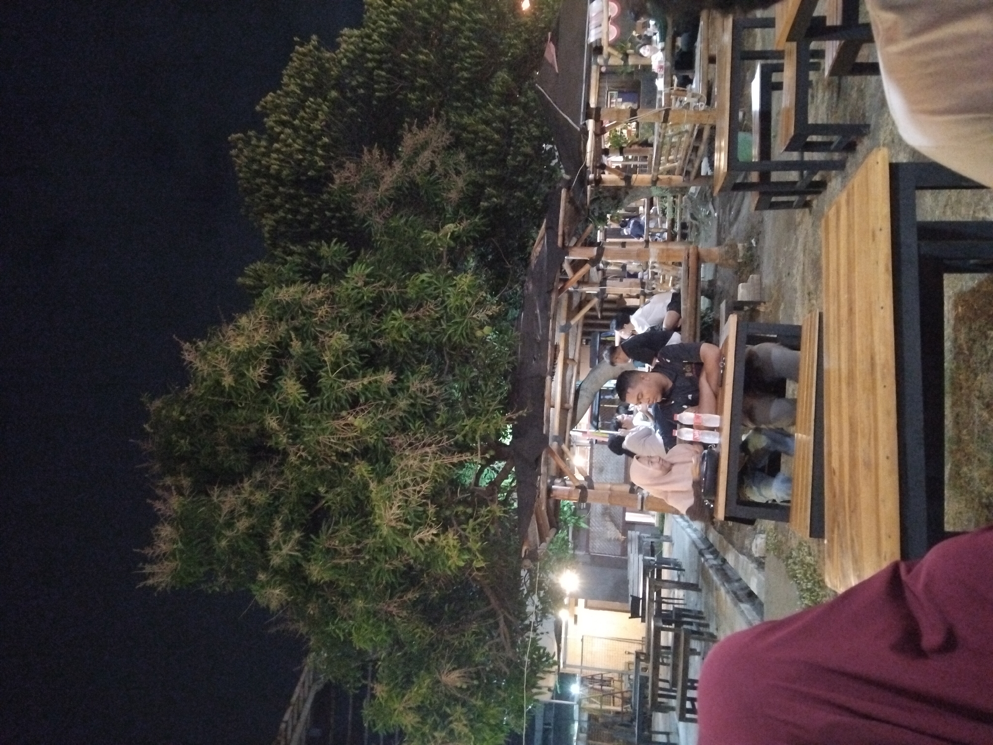 This is a night-time outdoor scene at a restaurant in Betro, Indonesia, possibly a seafood restaurant given the ambiance of similar establishments in the area. The foreground features a wooden table and benches. In the mid-ground, a couple is seated at a table, the woman wearing a hijab, and the man wearing a dark polo shirt. They are facing each other, suggesting interaction. Two water bottles are visible on their table. Behind them, the restaurant appears to be open-air with bamboo structures and wooden tables and benches, and other patrons are visible in the background. Large trees with green foliage and some flowering branches are prominent in the scene, illuminated by ambient lighting. The overall atmosphere is casual and relaxed.