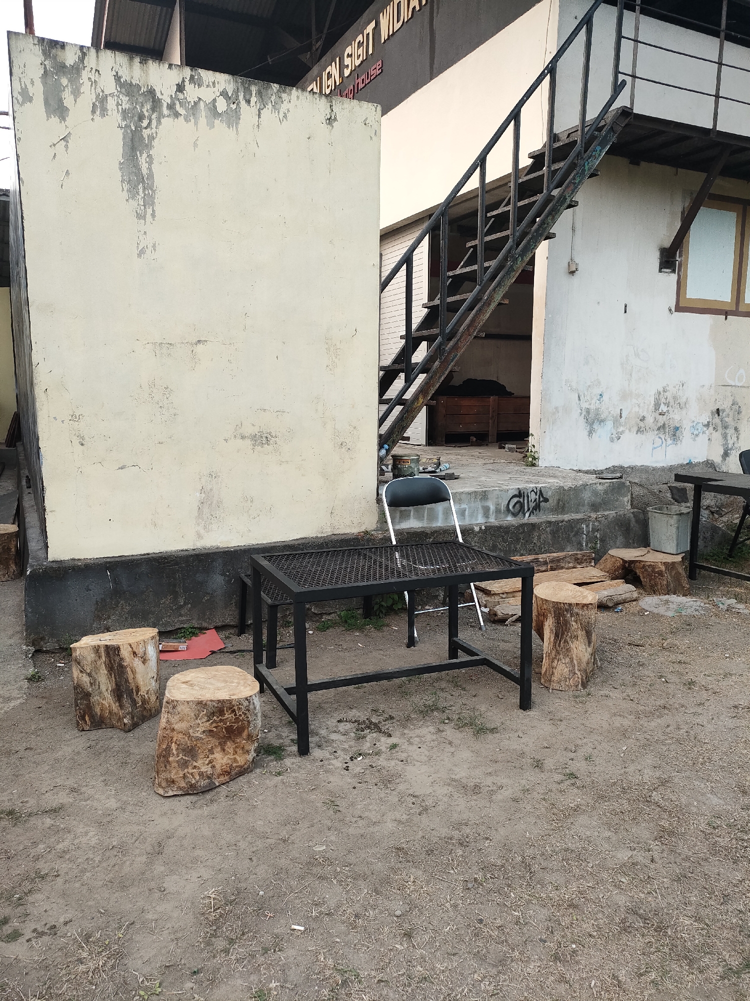 The image shows an outdoor seating area in Banguntapan, Indonesia. The scene features a metal barbecue grill with a black metal frame, positioned on a dirt ground. Around the grill, there are three wooden log stools. To the left of the grill, a white and black metal chair is placed. In the background, a building with a weathered white facade and a metal staircase leading to an upper level is visible. The building has a dark-colored roof and some graffiti on its walls. The ground is composed of dry soil and scattered dry grass and debris. The overall impression is of a casual, possibly rustic, outdoor dining or gathering space.