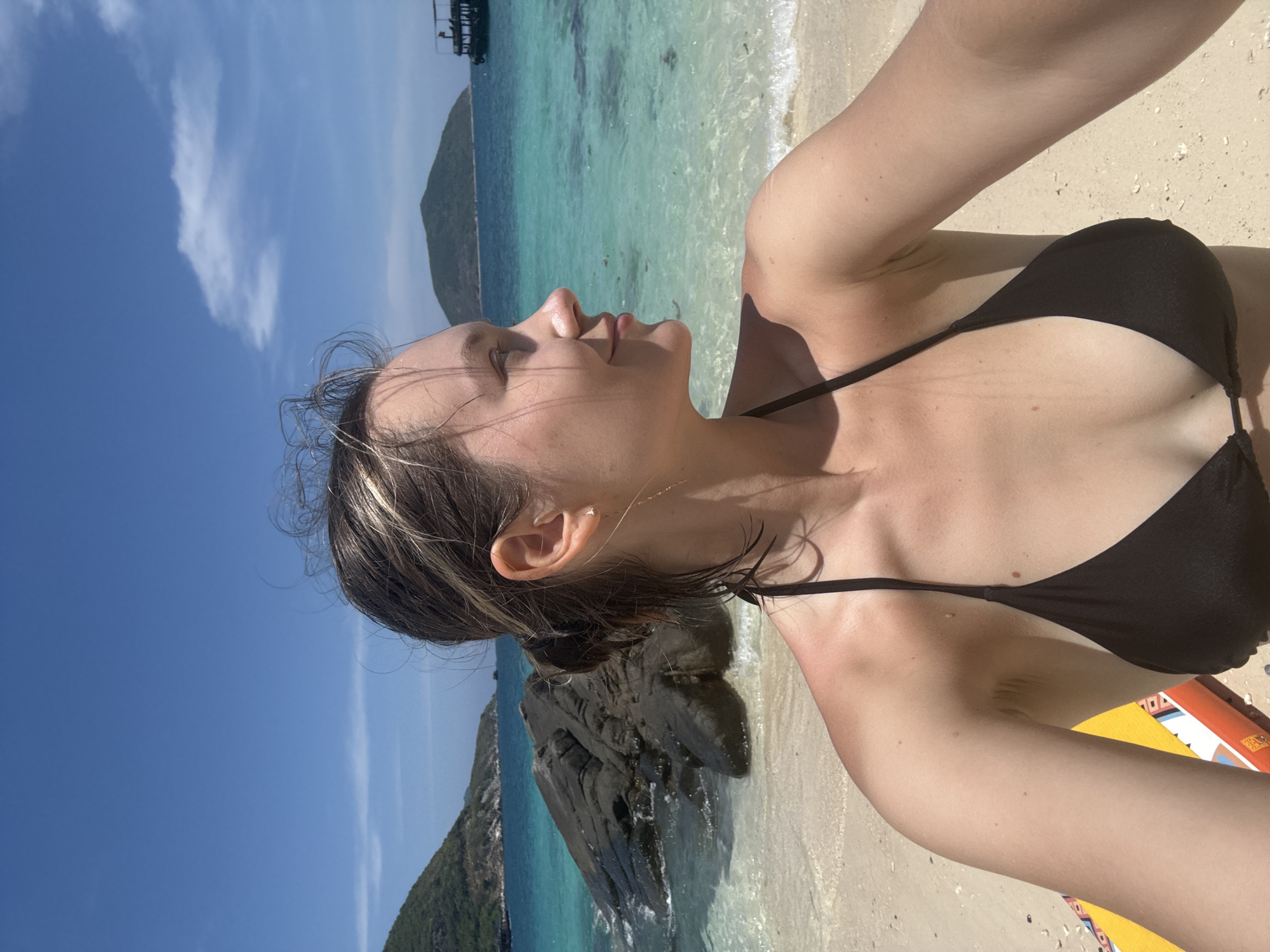 A woman with fair skin, wearing a black bikini, is lying on a sandy beach. She is looking upwards towards the sky. The background shows clear turquoise water, a rocky outcrop, and a green, hilly island. A boat is partially visible in the distance. The location is suggested to be Nha Trang Ward, Vietnam.