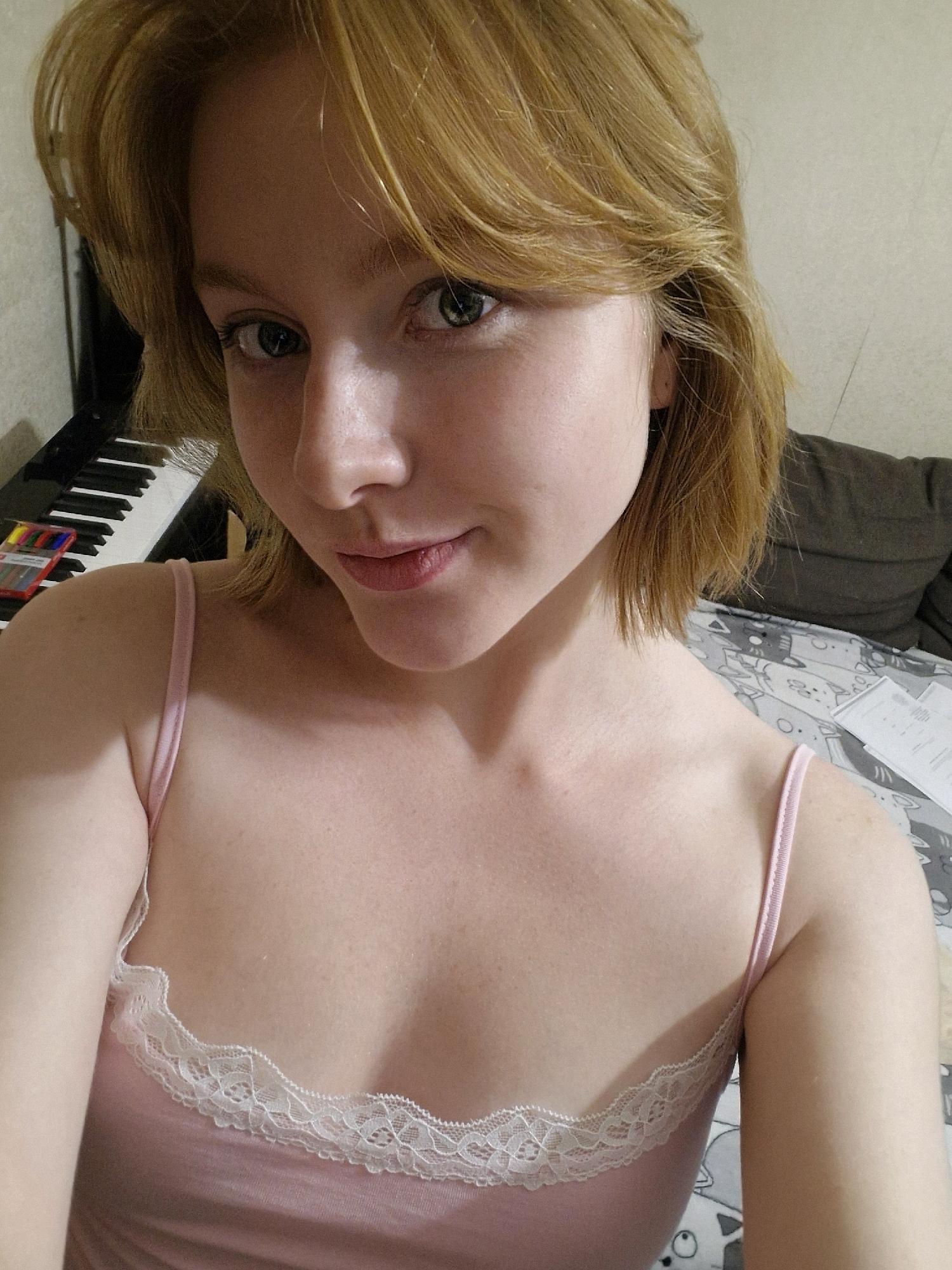 The image shows a close-up, waist-up portrait of a young woman with blonde hair and green eyes, wearing a pink camisole with lace trim. She is indoors, with a keyboard and some colorful markers visible in the background on the left, and what appears to be a bed or sofa with patterned sheets on the right. There is no indication of Yekaterinburg, Russia in the image. The lighting is soft and natural, suggesting it might be daytime.