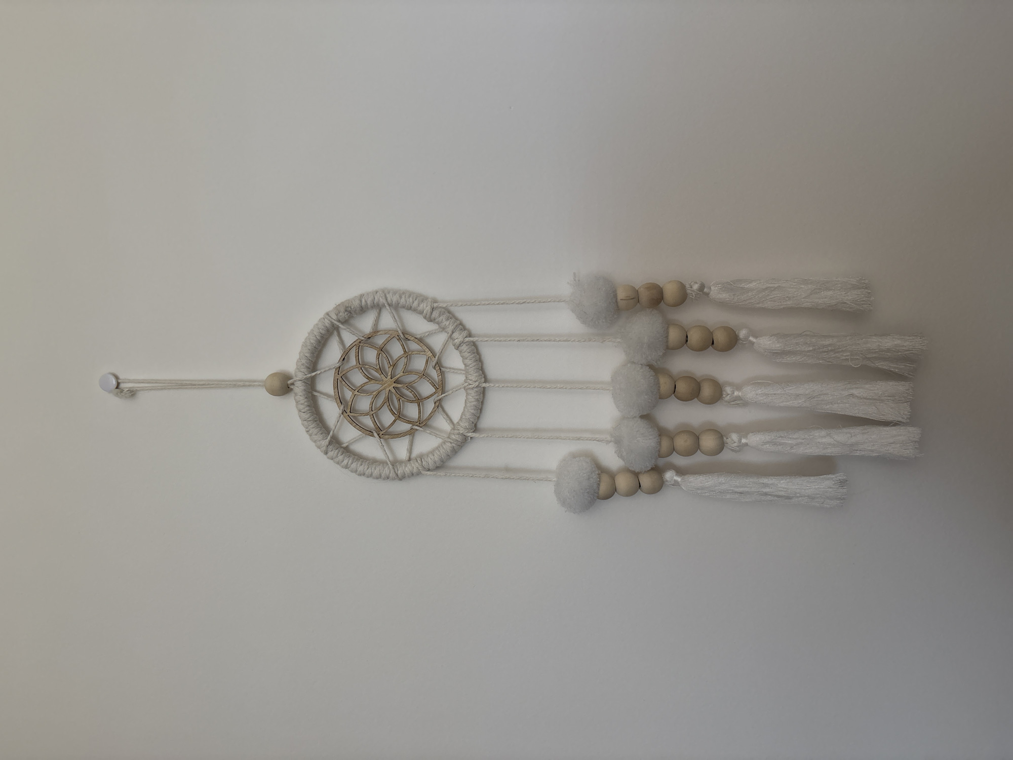 A cream-colored dreamcatcher is hanging on a plain white wall. The dreamcatcher has a circular woven frame with a wooden disc in the center, featuring a geometric flower pattern. Four cream-colored strings hang down from the frame, each adorned with wooden beads and topped with a white fluffy pom-pom. At the bottom of each string are white tassels. The scene is devoid of any people or recognizable urban elements. The image appears to be a product shot taken in an indoor setting. No specific location in the city of Liberec, Czechia can be identified from the provided image.