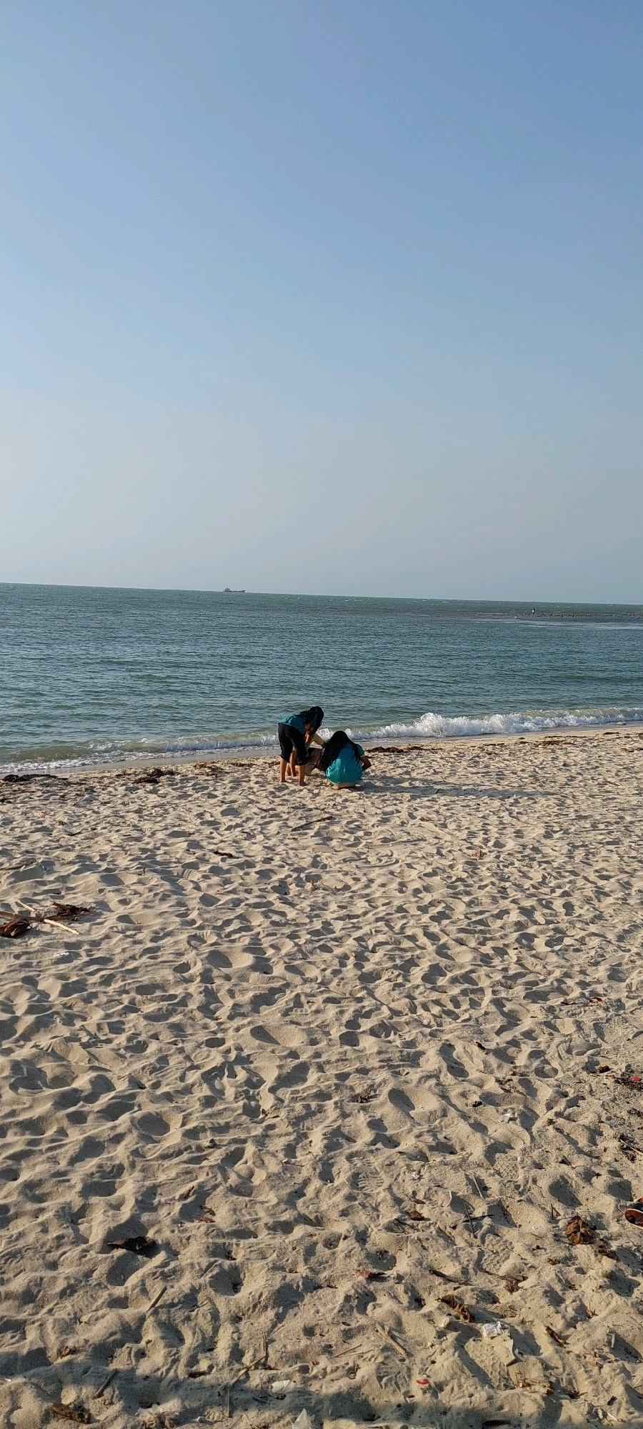 The image depicts a beach scene in Karanggondang, Indonesia. The sky is a clear, light blue, with a faint haze near the horizon. The sea is a calm, greenish-blue, with gentle waves lapping at the shore. The beach itself is covered in light-colored sand, marked with numerous footprints and small debris such as dried leaves and twigs.
In the middle ground, two individuals are interacting on the sand near the water's edge. One person, wearing a dark top and dark shorts, is bent over, appearing to engage with something on the ground. The second person, wearing a teal-colored garment, is crouched down next to the first individual, also focused on the ground. Their actions suggest they might be playing in the sand or examining something.
Further out in the sea, a small, dark ship is visible on the horizon, indicating maritime activity. The overall environment is serene and appears to be a sunny day.