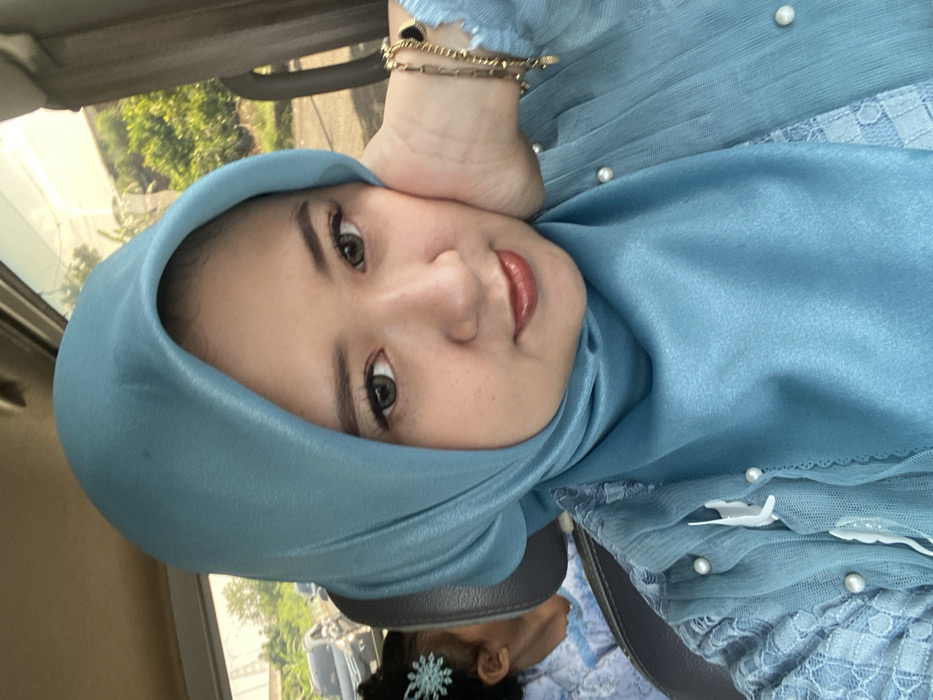 An image captured from within a vehicle depicts an adult female in the foreground, with partial views of another individual and an outdoor environment in the background. The primary subject, of presumed East Asian descent, occupies the majority of the frame, facing the camera directly. She wears a solid teal hijab and a matching teal top featuring patterned lace and white pearl details on the bodice. Her left hand is positioned against her cheek, supporting her head. A gold-colored bracelet, composed of multiple delicate chains and a small dark pendant, is visible on her left wrist. Her eyes are light-colored, possibly from contact lenses, and her lips are painted with a reddish-brown hue. Behind the primary subject and to her left, the top of a child's head is visible, adorned with a light blue, snowflake-shaped hair clip. This child is seated in what appears to be a car seat. Through the vehicle's windows, a sunlit outdoor scene is observable, including a road on the left with at least one dark-colored vehicle and distant utility poles, alongside dense green foliage on both sides. The scene is situated within a vehicle in the city of Kebonagung, Indonesia.