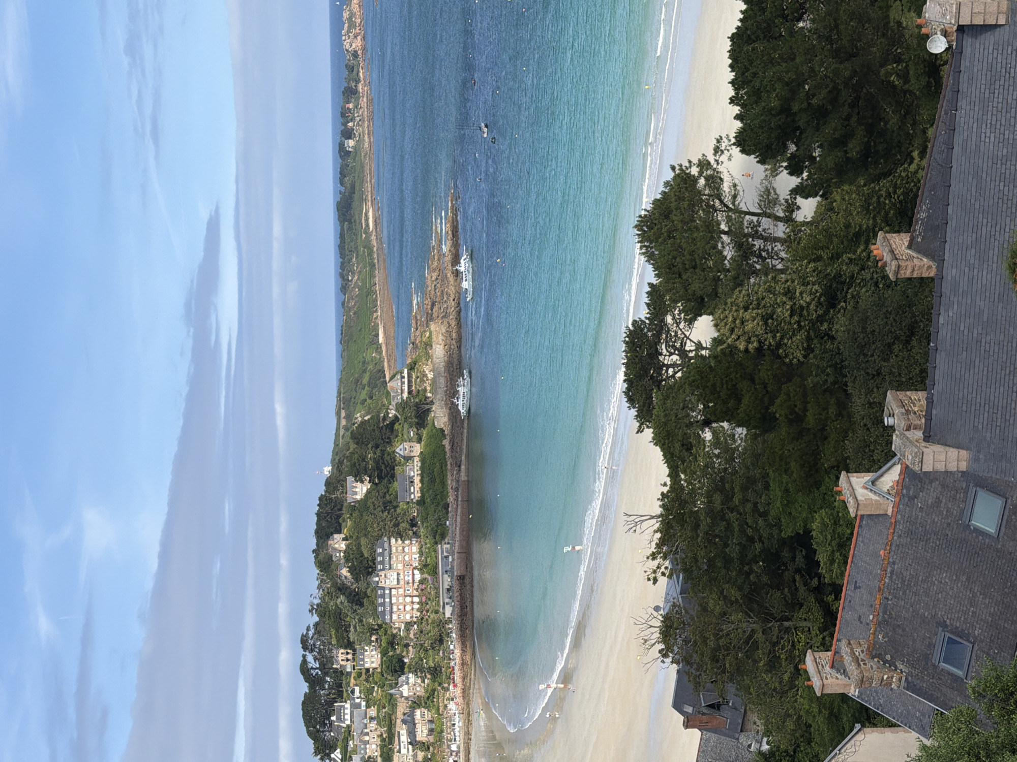 The image depicts a scenic coastal view of La Clarté, France. The scene is dominated by a tranquil turquoise sea meeting a sandy beach, with lush green trees and houses dotting the hillside in the background. The sky is a clear blue with wispy white clouds, suggesting a pleasant, sunny day. Several boats are visible in the water, some anchored and others appearing to be in motion. Small figures of people can be seen on the beach, enjoying the seaside. The foreground features the rooftops of houses, with chimneys and dormer windows, hinting at a residential area overlooking the coast. The overall impression is one of a peaceful, idyllic vacation spot. The time of day appears to be late morning or early afternoon, given the brightness and angle of the sun.