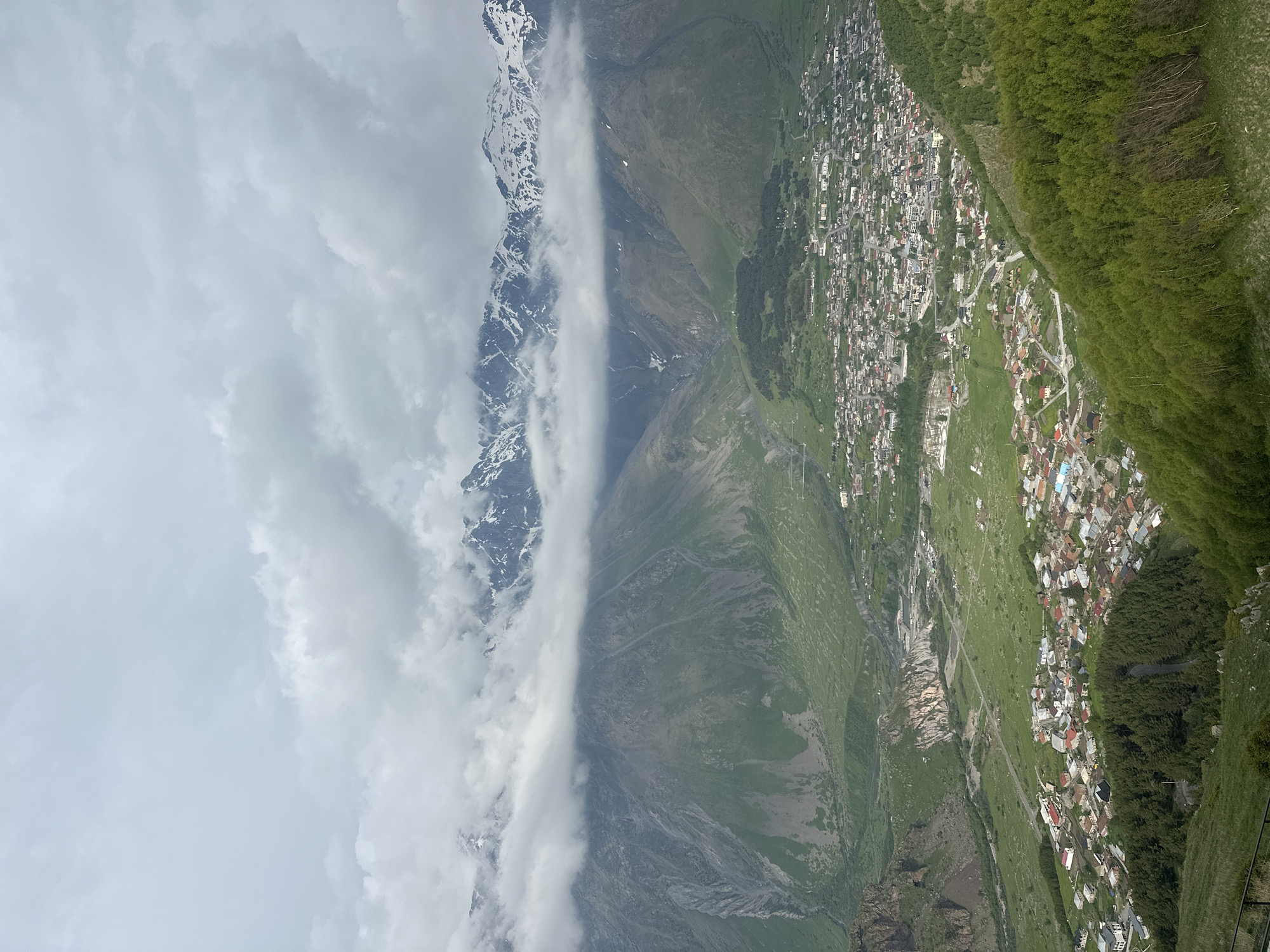 The image captures a panoramic view of the city of Stepantsminda, Georgia, nestled within a lush green valley. Dominating the background are the majestic, snow-capped peaks of the Greater Caucasus Mountains, partially obscured by dramatic, low-hanging clouds. The town itself is spread out below, a tapestry of red-tiled roofs and various structures, hinting at a dense urban fabric. Verdant hillsides slope down towards the valley floor, dotted with trees and interspersed with the urban development. A winding road is visible, leading into and through the town, suggesting connectivity. The overall impression is one of a remote, picturesque mountain town under an overcast sky.