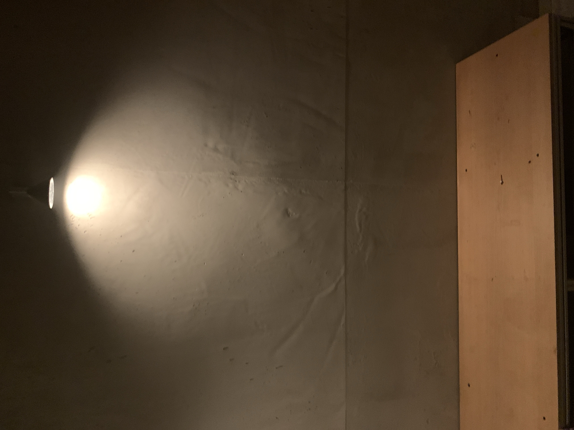 The image shows an interior space. The walls are painted in a light, neutral tone and appear somewhat textured. A bright light source, possibly a lamp or flashlight, is positioned to the left, casting a cone of illumination across the wall. To the right, a wooden panel or door is visible. No people or other animate subjects are present. The environment appears to be a storage room, basement, or utility space. The location is not discernible from the image.