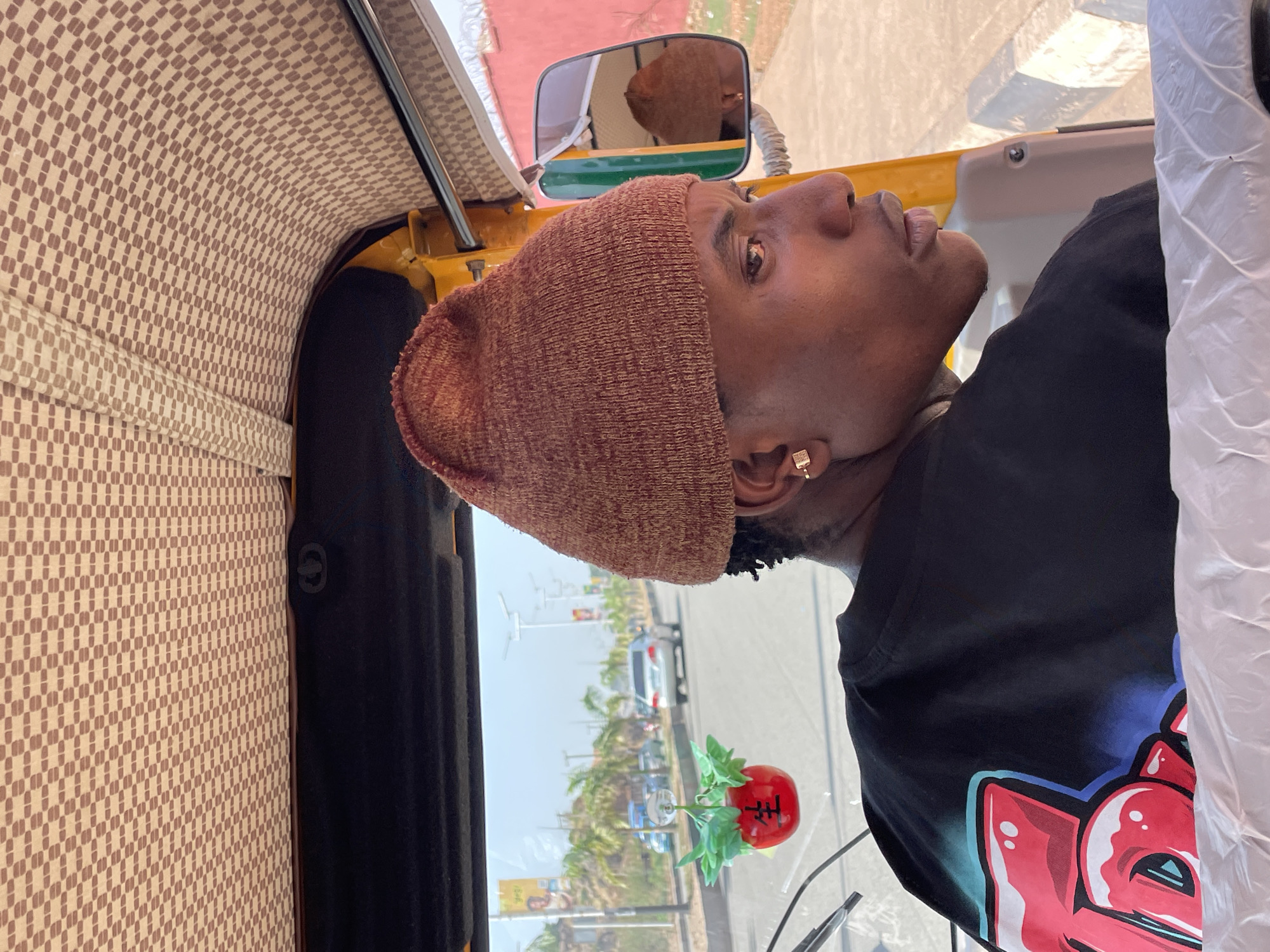 A medium-shot image shows a person in the driver's seat of an auto-rickshaw, likely taken within the city of Jos, Nigeria, based on environmental context. The individual, a Black male, is wearing a brown knit hat and a black t-shirt with a graphic design. He has a gold earring and is looking out the front of the vehicle. The auto-rickshaw features a brown and beige patterned interior roof, and a side mirror reflects the driver. Outside, palm trees, vehicles, and buildings are visible through the front windshield. A small, decorative red apple with lettering is attached to the dashboard. The background suggests a city street during daylight hours.