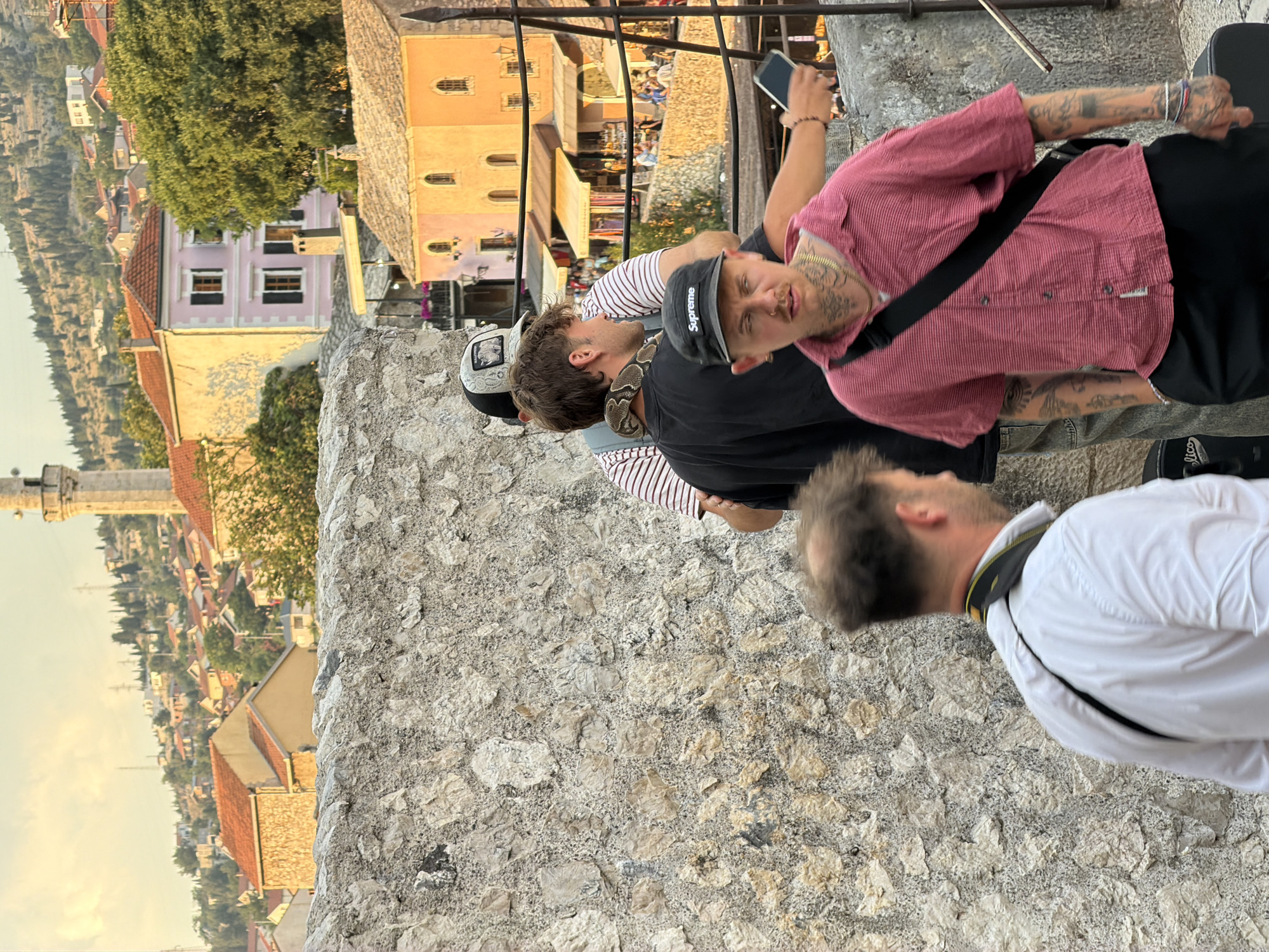 The image captures a scene in Mostar, Bosnia and Herzegovina, at what appears to be golden hour, given the warm, soft light bathing the landscape. The primary focus is on a group of young men, with at least three clearly visible. One man in the foreground, wearing a pink striped shirt and a black cross-body bag, has a snake draped around his neck and shoulders. His face is visible, and he has numerous tattoos. He is looking towards the camera. To his left, another man wearing a black t-shirt and a black "Supreme" baseball cap is also looking towards the camera. A third man, partially obscured, wearing a striped t-shirt, is also present.
Behind the individuals, a textured stone wall forms the foreground. Beyond this, the cityscape of Mostar unfolds. Traditional Balkan architecture with terracotta roofs and pastel-colored buildings are visible, set against a backdrop of a lush green hillside. In the distance, the iconic Stari Most bridge or a similar structure might be implied by the context of Mostar, though it is not distinctly visible in this frame. The setting suggests they are on a high vantage point, perhaps a fortress wall or a viewpoint overlooking the city.
The mood appears casual and perhaps adventurous, with the snake being a prominent and unusual element. The overall impression is a candid moment captured during a visit to the historic city.