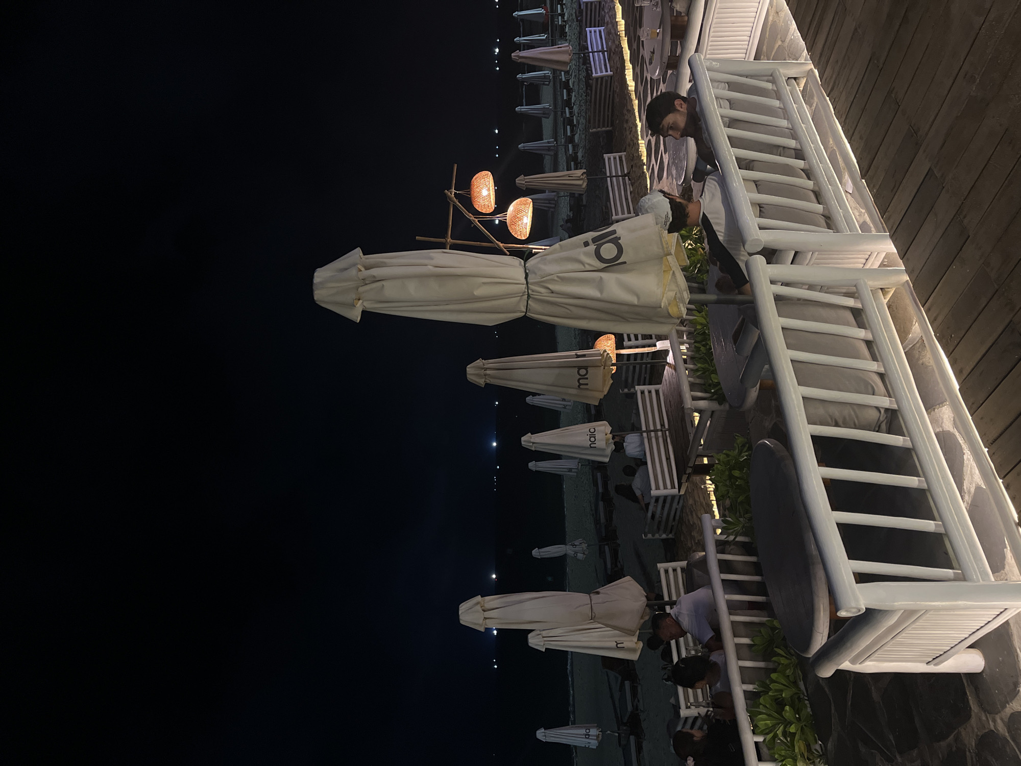 The image shows a nighttime outdoor scene along a coastline, likely in Đà Nẵng, Vietnam. A partially visible dark sky occupies the left side of the frame, contrasted by artificial lighting illuminating the beach area. Numerous closed beach umbrellas are arranged in rows. Wicker-style hanging lanterns provide warm light near the center of the image.
On the right, a section of an elevated outdoor terrace area is visible. White-painted wooden lounge chairs with gray cushions are neatly arranged. People, possibly patrons, are sitting and standing near the chairs. Details on their actions and interactions are limited due to the image's perspective and darkness. The terrace floor appears to be made of dark-colored wooden planks.