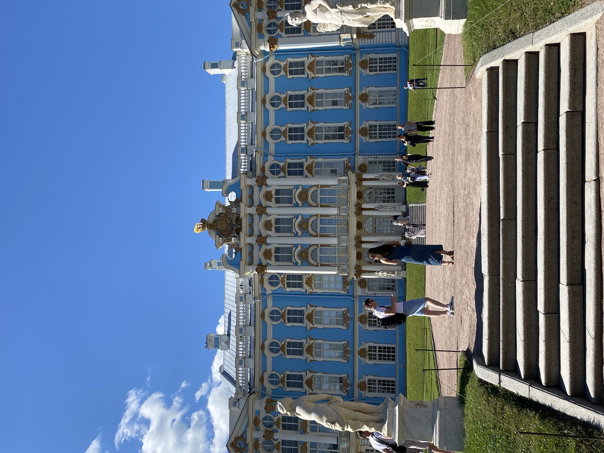 A bright, sunny day in Pushkin, Russia, is captured in this image, with the striking Catherine Palace as the central focus. The palace is a grand blue structure with white and gold ornate detailing and numerous windows. In the foreground, a wide set of stone stairs leads up towards a gravel path. Several people are visible on the path, some walking towards the palace, others engaged in conversation or looking at their phones. Two large marble statues stand on pedestals on either side of the path, facing the palace. Green grass surrounds the gravel area, and a clear blue sky with a few wispy clouds completes the scene. The overall impression is one of a pleasant, leisurely day at a historic landmark.