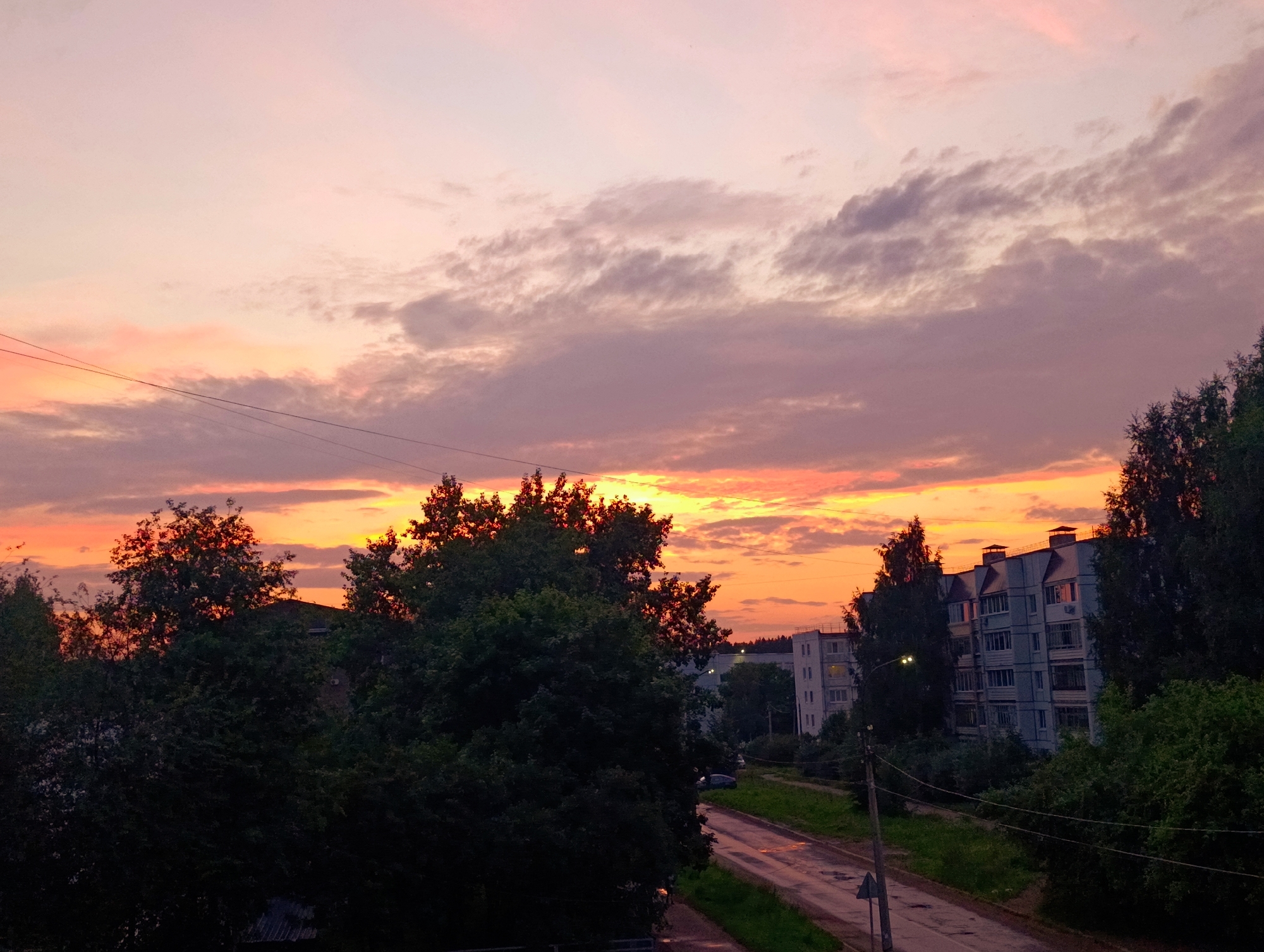 This is a wide-angle, eye-level, outdoor shot of a sunset over the city of Chaikovsky, Russia. The sky is a vibrant mix of pink, orange, and purple hues, with scattered clouds. In the foreground, silhouetted trees and some residential buildings are visible. A road runs diagonally through the scene, with some parked cars. The overall impression is one of a peaceful evening in a residential area.