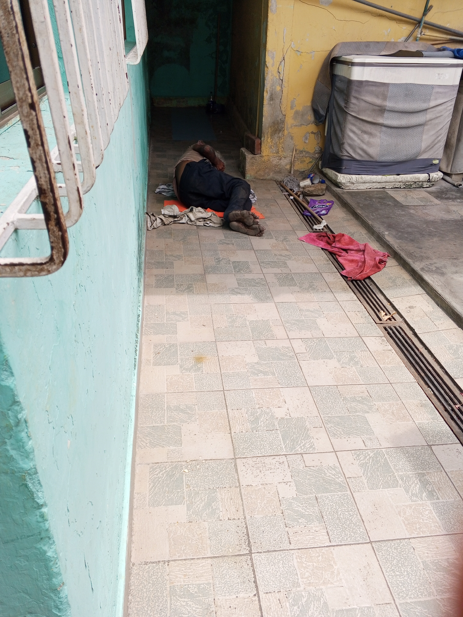 A man is sleeping on a tiled patio in Tinaquillo, Venezuela. He is lying on a makeshift bed of cloths and is wearing dark pants. His bare feet are visible. Next to him is a covered cooler, cleaning supplies, and a red cloth. The patio is bordered by a pale blue wall with a window and a yellow wall with peeling paint. A drainage ditch runs along the edge of the patio.
