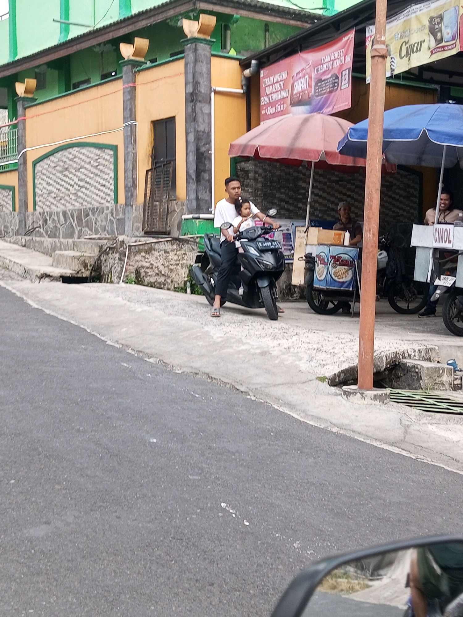 A man and a young child are on a scooter on a street in Krasak, Indonesia. The man, wearing a white t-shirt and dark pants, is driving the scooter with the child sitting in front of him. The child is wearing a light-colored shirt. They are in front of a building with an orange and green facade. There are several businesses with umbrellas set up on the sidewalk. One business has a sign that says "CIMOL". The street is paved and appears to be somewhat uneven. The image is taken from a low angle, possibly from another vehicle.