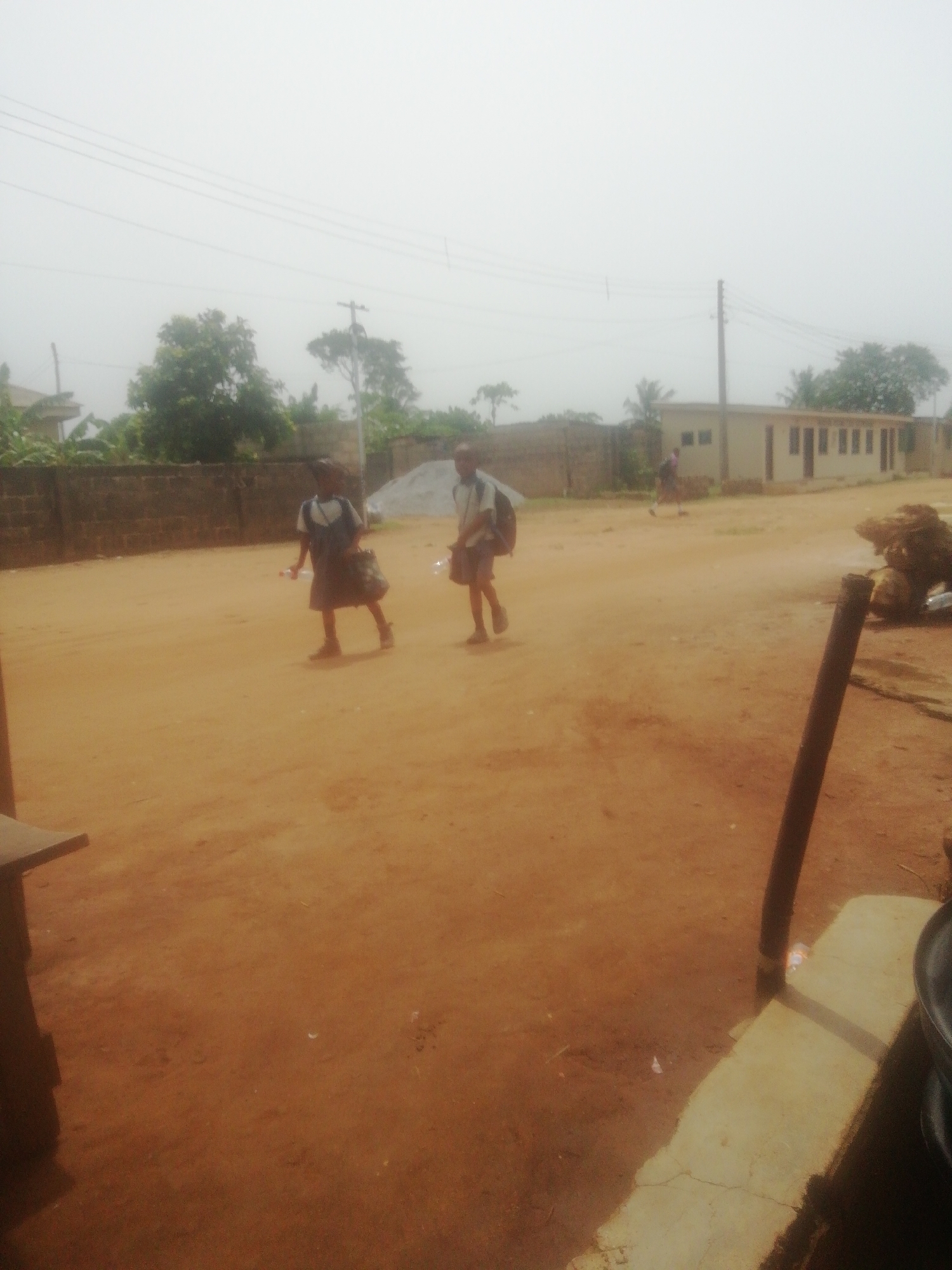 Two children in school uniforms are walking on a dirt road in what appears to be a village or rural area. A wall is visible on the left side of the road, and there's a building in the background on the right side. Trees and utility poles can be seen in the distance. Another person is further down the road near the building. Parts of structures and objects are visible in the foreground on either side of the image. The location is Unknown, Unknown.