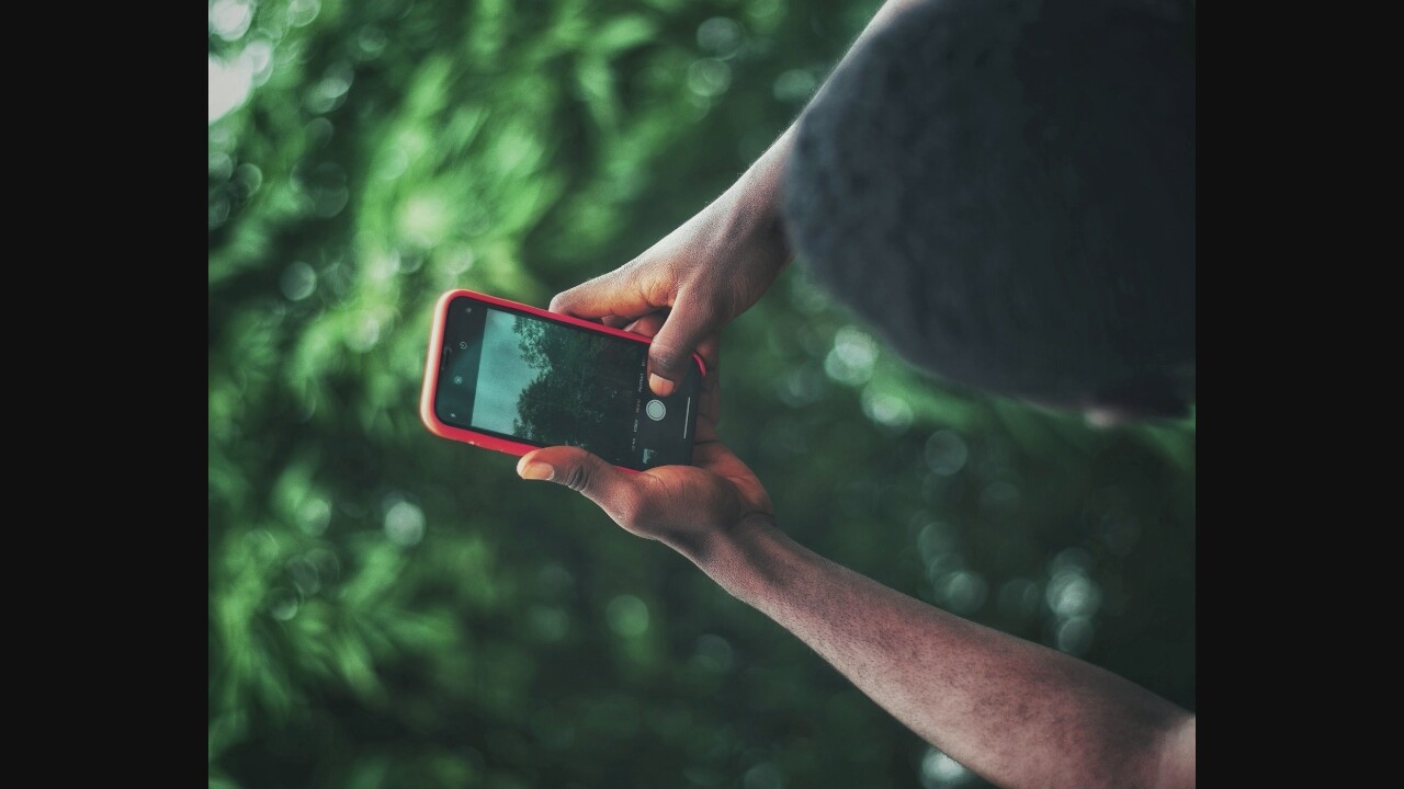 An over-the-shoulder medium shot captures a person's hands holding a smartphone with a red case. The phone displays a camera interface, and the screen shows a dimly lit forest scene with trees. The person's arms and hands, with visible dark skin and fine hairs, are positioned to hold and operate the phone. The background is a blur of lush green foliage, suggesting an outdoor, natural setting, possibly a park or wooded area within the city. The lighting is soft and diffused, contributing to a slightly moody atmosphere.