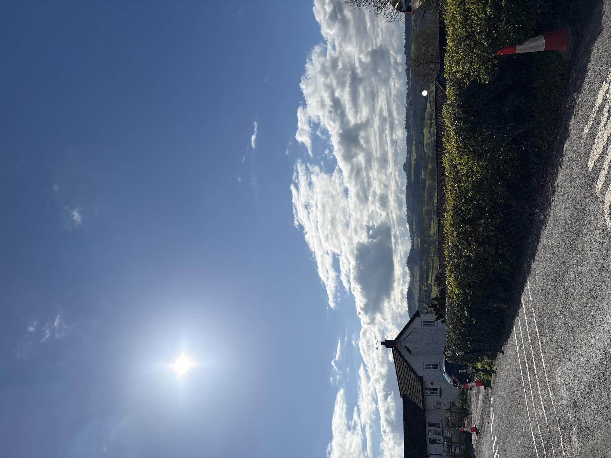 The image is a rotated view of a scene in Snailbeach, United Kingdom. It shows a clear, blue sky with cumulus clouds and the sun shining brightly. The sun is visible in the lower-left corner of the image. A white house with a dark roof can be seen in the lower center of the image. Adjacent to the house is a road, and beside the road is a dark green hedge and some tall grass. An orange and white road safety cone is by the hedge and road. In the distance, there are rolling green hills and mountains under the clouds.