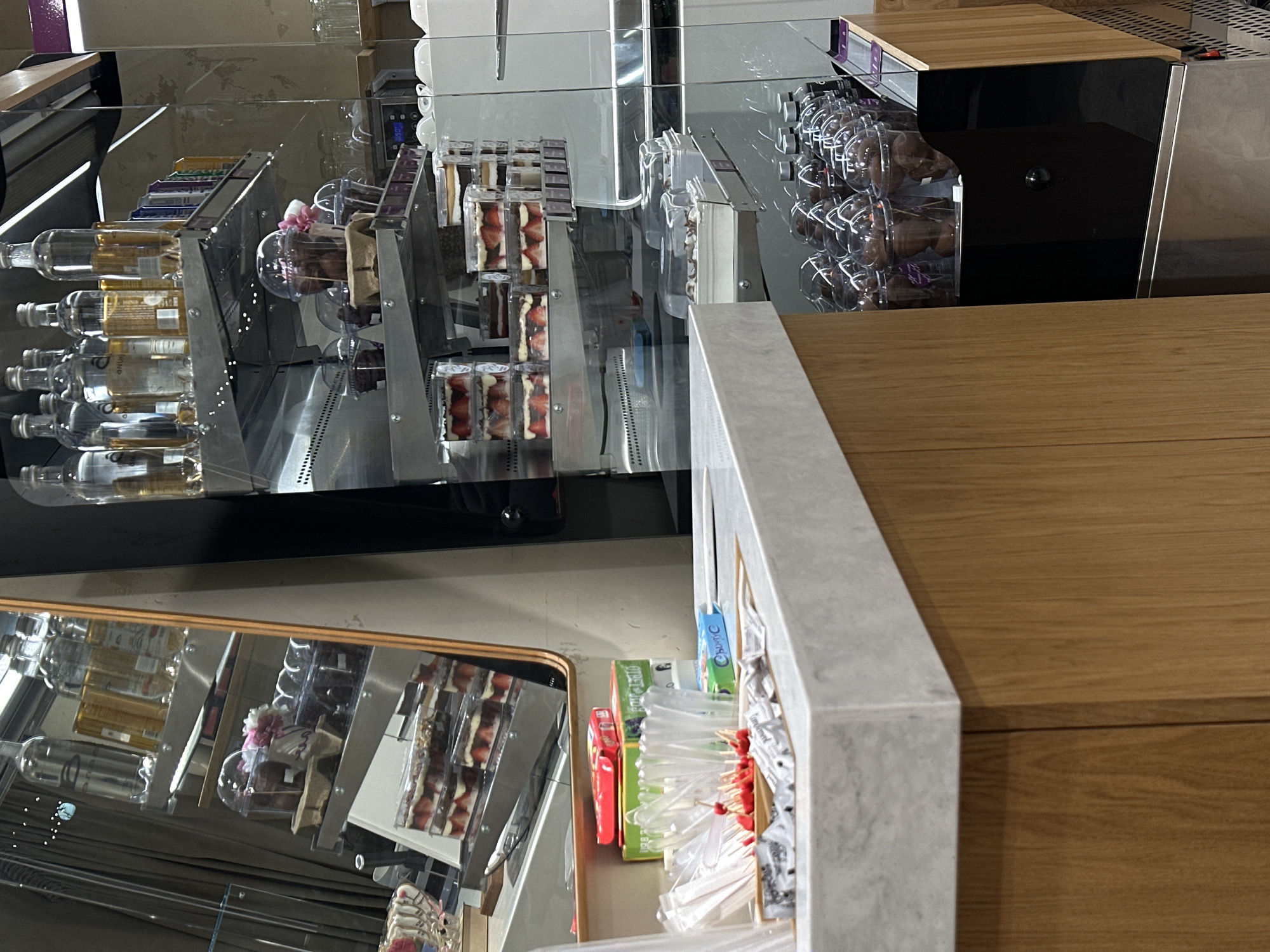 The image presents a commercial interior with reflections enhancing the visual complexity. The foreground features a wooden counter or shelf with a gray countertop trim, to the right of which are various packaged items, including what appear to be condiment packets and small boxes with colorful packaging.
Behind this countertop is a glass display case with multiple compartments, housing a variety of food items. There are rows of what appear to be bottles of liquids, possibly oils or syrups, and layered cakes or pastries, and chocolate desserts in individual portions.
The scene appears to be set in a modern cafe or gourmet food store, potentially in Rostov-on-Don, Russia. However, without further contextual clues, the location can only be inferred. The scene contains no people.