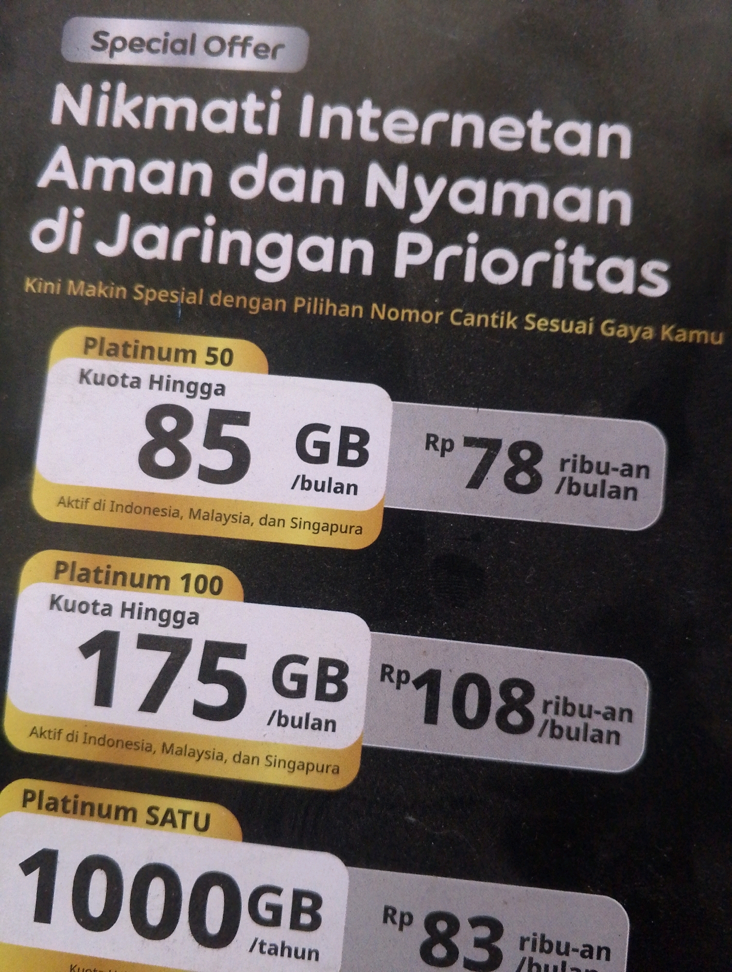 The image displays an advertisement for internet plans. The headline reads "Nikmati Internetan Aman dan Nyaman di Jaringan Prioritas" which translates to "Enjoy Safe and Comfortable Internet on a Priority Network." Below this, a smaller line states "Kini Makin Spesial dengan Pilihan Nomor Cantik Sesuai Gaya Kamu" meaning "Now Even More Special with a Choice of Beautiful Numbers to Suit Your Style."
The advertisement then lists three different internet packages:
1. **Platinum 50:** This plan offers "Kuota Hingga 85 GB /bulan" (Up to 85 GB per month) and is "Aktif di Indonesia, Malaysia, dan Singapura" (Active in Indonesia, Malaysia, and Singapore). The price is "Rp 78 ribu-an /bulan" (Rp 78 thousand per month).
2. **Platinum 100:** This plan offers "Kuota Hingga 175 GB /bulan" (Up to 175 GB per month). The price is "Rp 108 ribu-an /bulan" (Rp 108 thousand per month). It also states it is active in Indonesia, Malaysia, and Singapore.
3. **Platinum SATU:** This plan offers "1000 GB /tahun" (1000 GB per year). The price is "Rp 83 ribu-an /bulan" (Rp 83 thousand per month), although the duration of this price is not explicitly stated as per year.
The advertisement is presented on a dark background, likely a poster or a flyer, with bright yellow and white elements to highlight the different plans and pricing. The overall aesthetic is modern and promotional. There are no people visible in the image. The image is a close-up of the advertisement, focusing on the text and pricing details. The location is unknown as it is an advertisement and not a scene from a specific city.