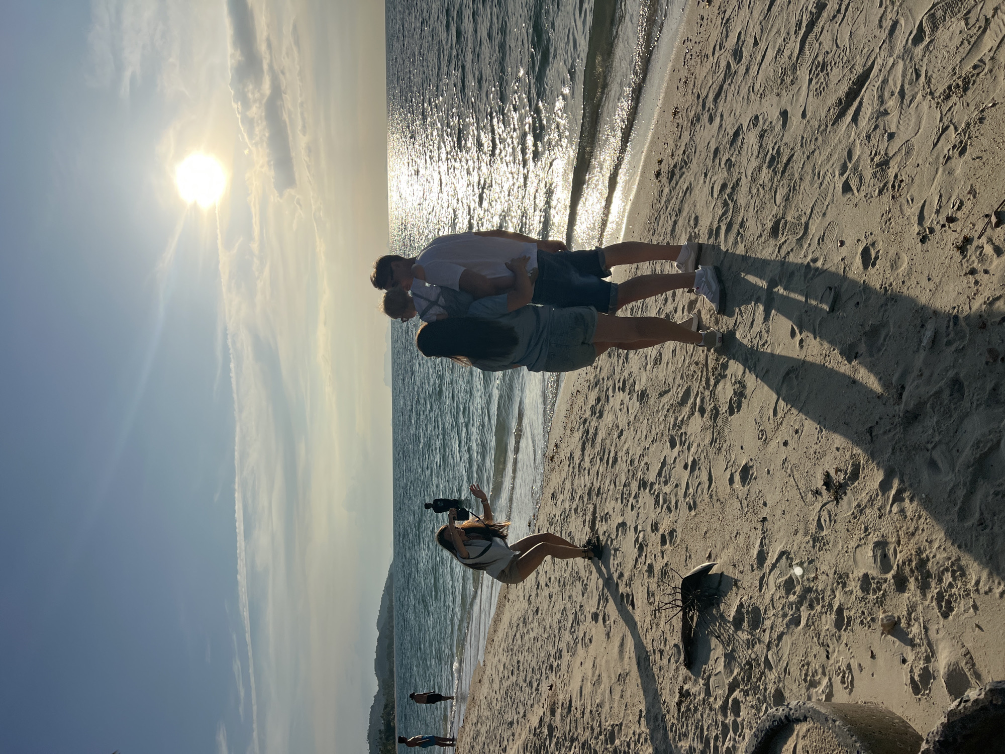 The image depicts a beach scene at Baan Tai, Thailand during daytime. Sunlight reflects off the water. A couple stands embraced at the shoreline, facing away from the camera. A photographer, seen in the foreground, holds a camera and appears to be taking their picture. Additional people are visible further down the beach. The beach sand shows numerous footprints and the shadows of the subjects. Debris, including what appears to be part of a tire and some driftwood, sits on the sand.