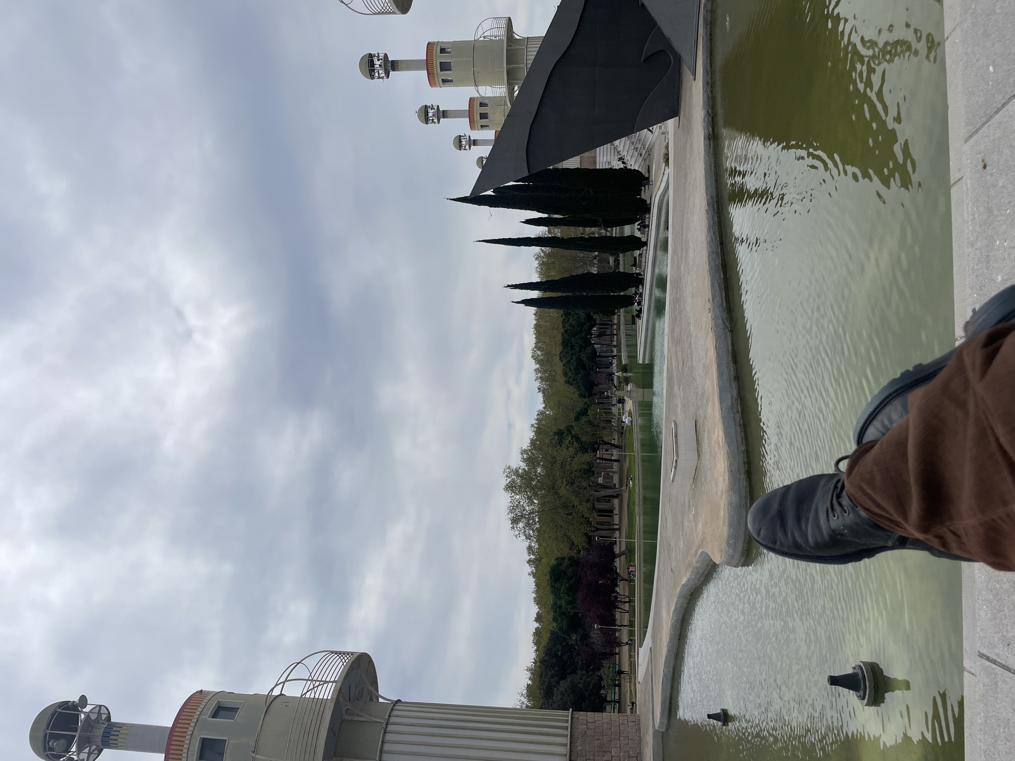 The image shows a partial view of the Magic Fountain of Montjuïc in Barcelona, Spain. A series of decorative towers, typical of the area, are visible in the background, along with tall, slender cypress trees and other greenery. The foreground features a section of the fountain's complex of pools, filled with greenish water. A person's leg, clad in brown trousers and a dark shoe, is placed at the edge of the fountain, suggesting an observer's perspective. The sky is overcast, casting a muted light over the scene.