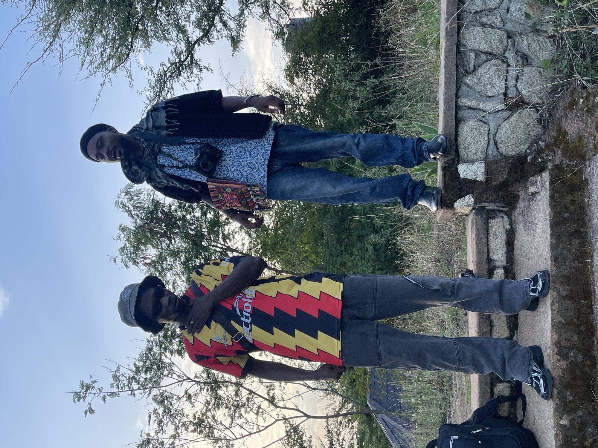 Two African men stand outdoors in Rayfield, Nigeria. One man wears a black and red zigzag-patterned shirt, grey pants, black shoes, and a grey bucket hat, with a black bag at his feet. The other wears a patterned blue shirt, black jacket, blue jeans, and black shoes, with a camera bag across his chest and a camera around his neck. They are standing on a stone structure with trees and bushes in the background. The sky is visible.