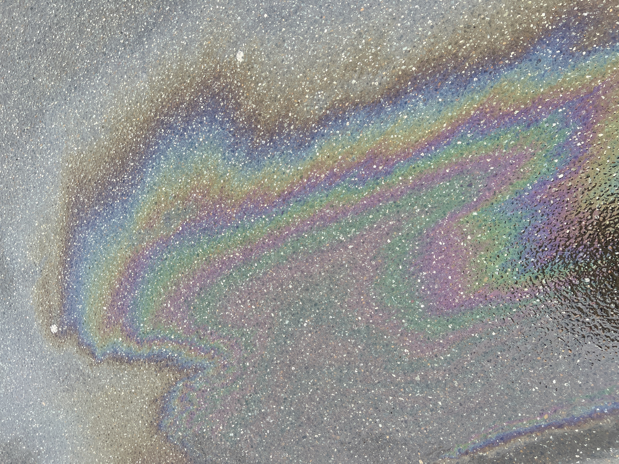 The image provides a close-up, high-angle view of a vibrant, iridescent oil or petrol slick on a coarse, paved ground surface, likely asphalt or concrete. The oil film covers a large, irregular area across the frame, showcasing a striking spectrum of colors, including blues, greens, yellows, oranges, reds, and purples. These colors swirl and blend in intricate patterns, characteristic of thin-film interference.
The underlying pavement exhibits a granular texture, composed of small, light-colored aggregate particles set within a darker matrix. Portions of the surface appear damp or wet, which contributes to the reflective quality and the vividness of the oil slick's colors. The overall bright illumination suggests that the photograph was taken during daylight hours.
The scene focuses exclusively on this natural phenomenon, with no people, vehicles, or specific environmental structures visible. The image captures an abstract and visually engaging detail found on a ground surface, likely in an urban setting such as a street or parking lot in Kazan, Russia. No visible text is present in the media file.