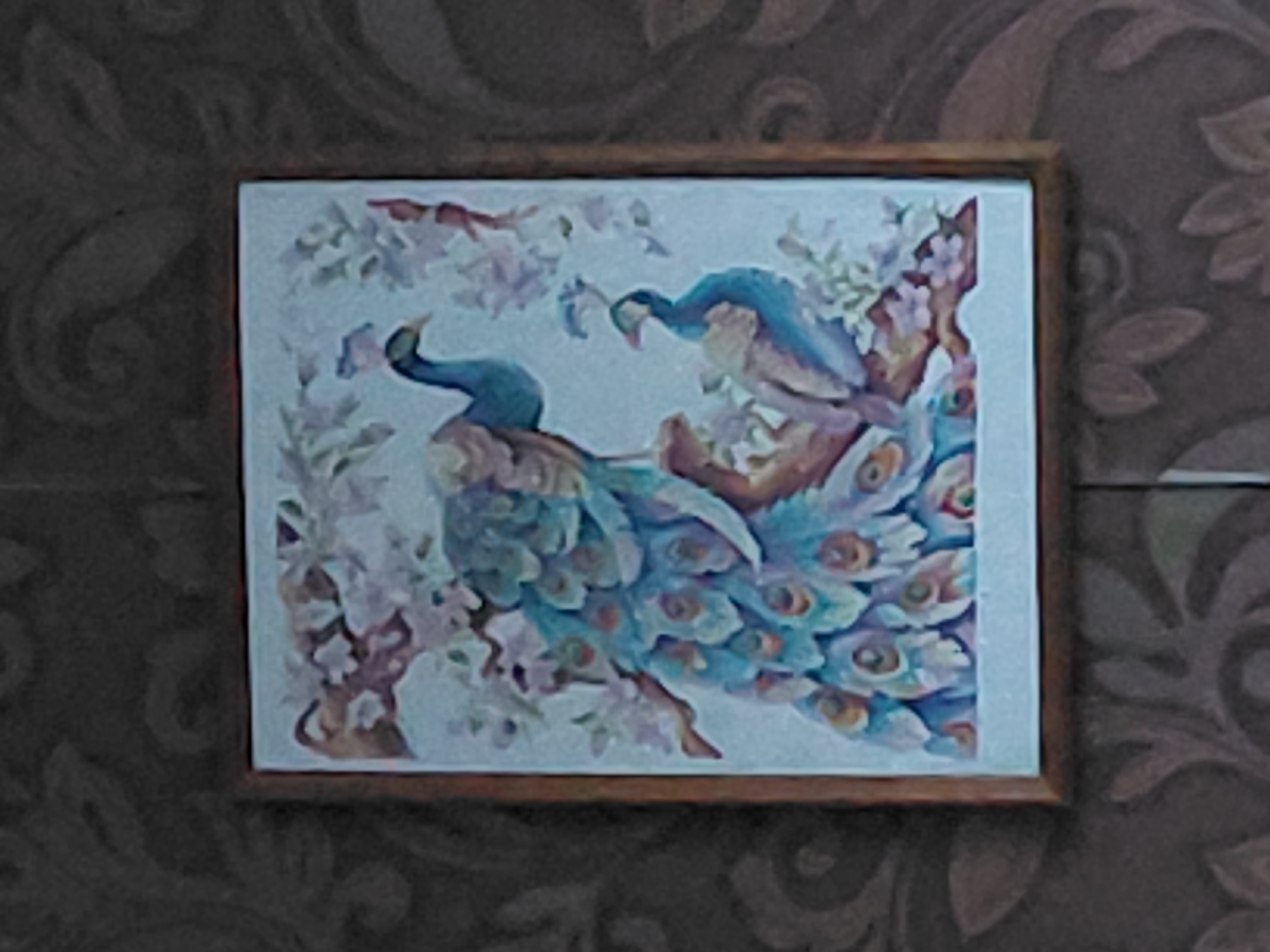 A framed painting featuring two peacocks is displayed on a textured, dark-colored wall with a floral pattern. The painting depicts one peacock with its tail fanned out, showcasing iridescent blues and greens with eye-like spots, perched on a branch. A second, smaller peacock is positioned below and to the right, also on a branch, with its neck curved. Delicate pink and purple flowers are interspersed throughout the branches and around the birds. The painting is framed by a simple, light-brown border. The overall lighting is dim, with the painting being the primary illuminated object. The image is taken from a frontal perspective.