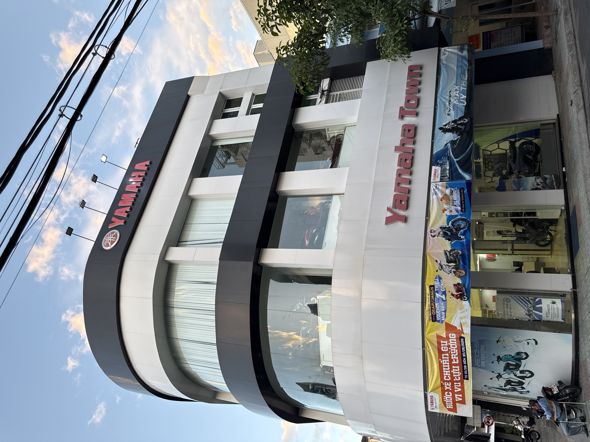 The image shows a Yamaha Town dealership in Nha Trang, Vietnam. The dealership is a multi-story building with a curved facade. The upper floors have large windows displaying motorcycles. A large red "Yamaha Town" sign is prominently displayed on the building's facade. Below the sign, a large banner advertises a promotion for Yamaha motorcycles. On the ground floor, the entrance to the dealership is visible, with motorcycles displayed inside. In front of the dealership, a man is sitting on a chair next to a motorcycle. The sky is blue with some clouds. The street in front of the dealership is paved, and there are trees and power lines visible. The building appears to be modern and well-maintained. The overall impression is of a busy commercial area in Vietnam.