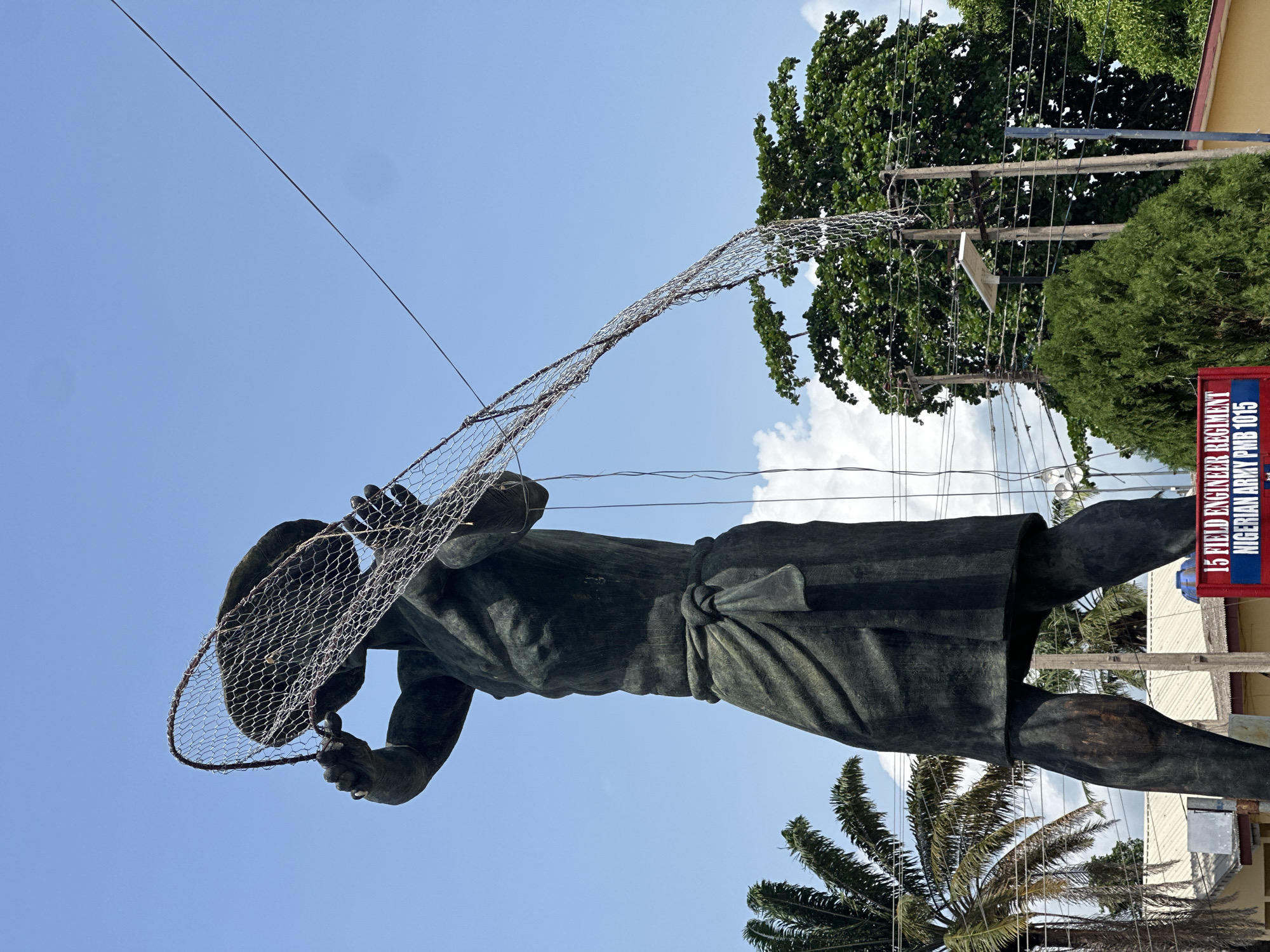 A large, dark-colored statue of a human figure is prominently featured, captured from a low-angle perspective against a bright, clear blue sky with a few scattered white clouds. The statue, likely crafted from bronze or a similar material, depicts a person wearing a simple garment, possibly a loincloth or wrap, tied around the waist. The figure's arms are raised, holding a large, wide-meshed net, reminiscent of a fishing net, which is spread open, suggesting an action like casting or displaying it.
Lush green foliage from several trees, including the distinctive fronds of a palm tree, frames the lower and right portions of the background. On the upper right, multiple utility poles crisscrossed with numerous electrical wires are visible, set against the backdrop of dense green tree canopy.
A notable detail is a partially visible red and white sign on the far right, bearing white text that reads: "15 FIELD ENGINEER REGIMENT NIGERIAN ARMY PMB 1015". This text strongly indicates a military presence and confirms the location's association with the Nigerian Army, aligning with the context of Badagry, Nigeria. The bright lighting and clear sky suggest the photograph was taken during sunny daylight hours. The overall impression is of an imposing public monument in a tropical or subtropical setting.