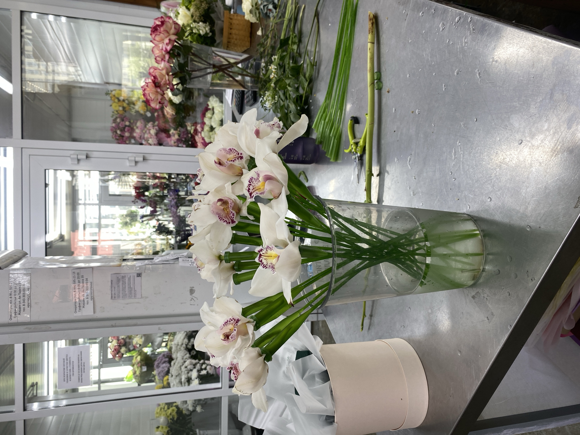Here is a detailed description of the image:
**Scene Overview:** The image appears to be taken inside a flower shop, likely in Yekaterinburg, Russia, based on the Cyrillic text visible in the background. A metal countertop dominates the foreground, displaying a vase of white orchids and various floral arrangement supplies. The background shows a window display of other floral arrangements and the shop's interior.
**Foreground Details:**
* **Vase of Orchids:** A clear glass vase holds several stems of white orchids. The flowers have distinctive purple/pink speckles in their centers. The stems are submerged in water.
* **Flower Box:** Near the orchids, there is a tan-colored cylindrical flower box, filled with white tissue or wrapping paper.
* **Floral Supplies:** On the right side of the countertop, there are green floral stems, shears, and other floral tools.
**Background Details:**
* **Window Display:** The window provides a view of the interior of the flower shop. Numerous floral arrangements in varying colors (roses, lilies, etc.) are displayed.
* **Cyrillic Text:** A sign with Cyrillic text is visible near the window, possibly shop information and contact numbers. The text indicates that the store is located on "Ak. Parina 39" street.
* **Shop Interior:** The interior shows more floral displays and the general layout of the flower shop.
**Location:** Flower Shop in Yekaterinburg, Russia, located on "Ak. Parina 39" street.
**Overall Impression:** The image seems to be a shot of a florist preparing or displaying orchids in a shop setting.