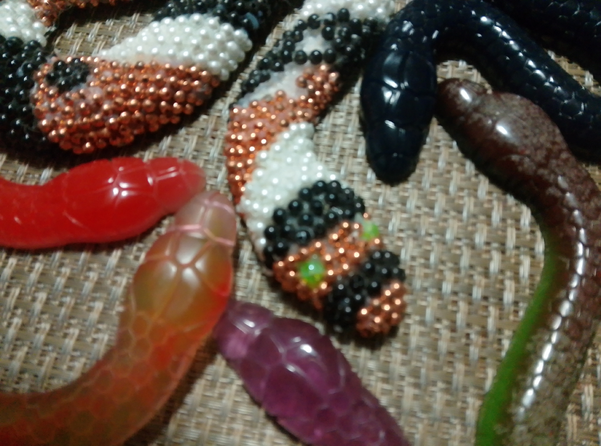 This image is a close-up, top-down view of several snake-shaped candy or toy items arranged on a textured, woven surface. The items are made of translucent, brightly colored plastic and appear to be arranged in a pile. From left to right, there is a red snake, followed by an orange-to-yellow translucent snake, a purple translucent snake, and then a black plastic snake. Further to the right and slightly behind the black snake is a dark brown, segmented snake. In the upper portion of the image, partially obscured, is a snake-like object covered in small, shiny beads, predominantly black and white, with some copper-colored beads and a few green bead "eyes." The surface they are on is a neutral, light brown woven material, possibly a placemat or fabric. There are no people or discernable environmental features that would suggest a specific location within the city of Wedel, Germany.