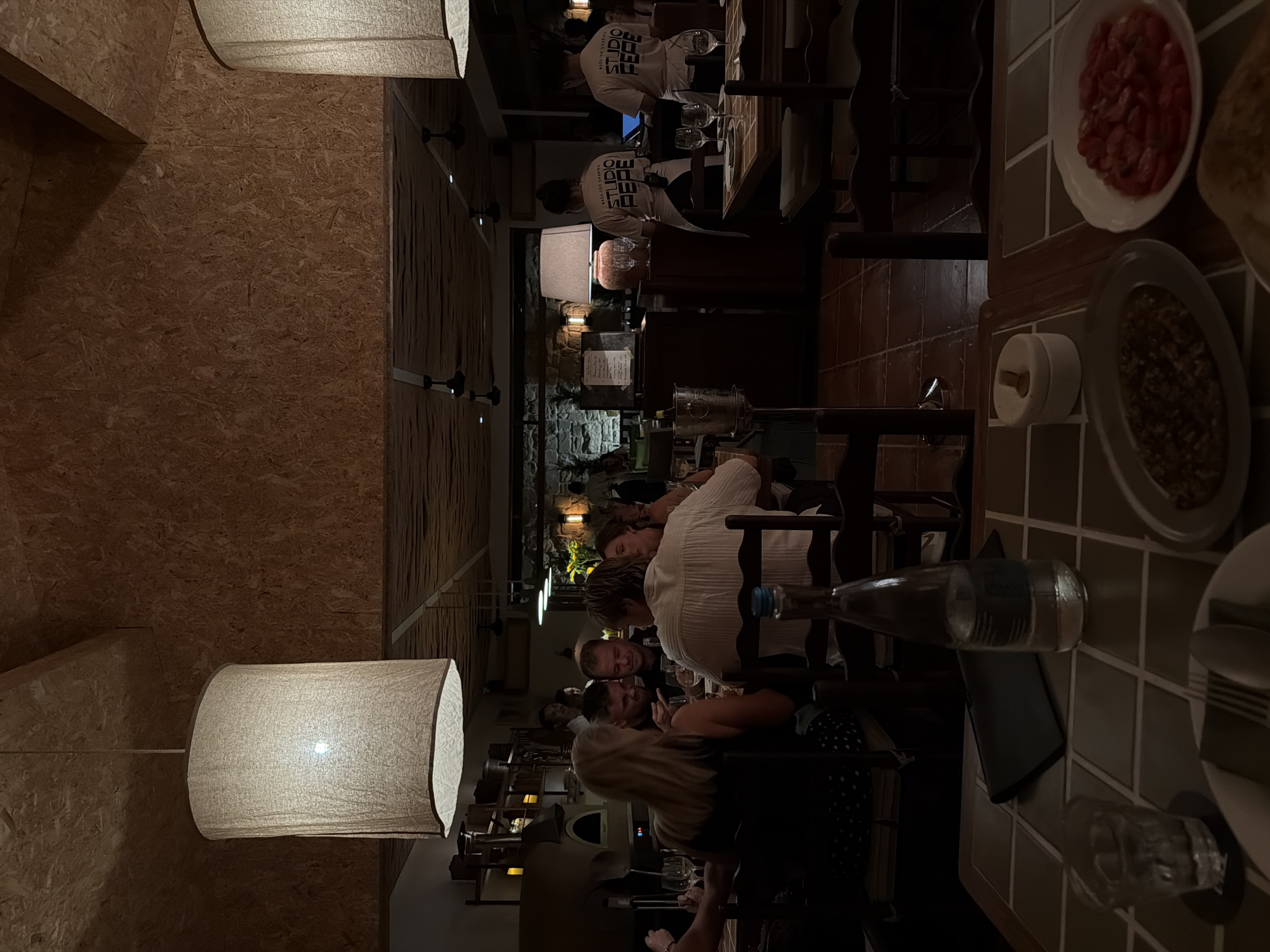 The image depicts a dimly lit interior of a restaurant, likely in North Kuta, Indonesia, at nighttime. The setting features wooden paneled walls and tables covered with a tiled surface. Several people are seated at tables, engaged in conversation and dining. The atmosphere appears relaxed and social, with warm lighting emanating from pendant lamps and wall sconces.
In the foreground, a large, cylindrical pendant lamp with a textured shade hangs from the ceiling, its light illuminating the immediate area. On the tables, various objects are visible, including plates of food, wine glasses, and a bottle of water. One plate contains sliced red tomatoes, while another holds a portion of lentils or a similar grain. A small, round white object with a pointed top sits on the tiled surface near the plates.
Further back, a group of individuals are seated together. One person, wearing a white t-shirt with "FF STUDIO" printed on it, is visible in the background. The people in the foreground appear to be in conversation, with some smiling and gesturing. The overall scene suggests a casual dining experience. The lighting creates a cozy ambiance, characteristic of an evening meal. No specific weather conditions are discernible from the indoor setting.