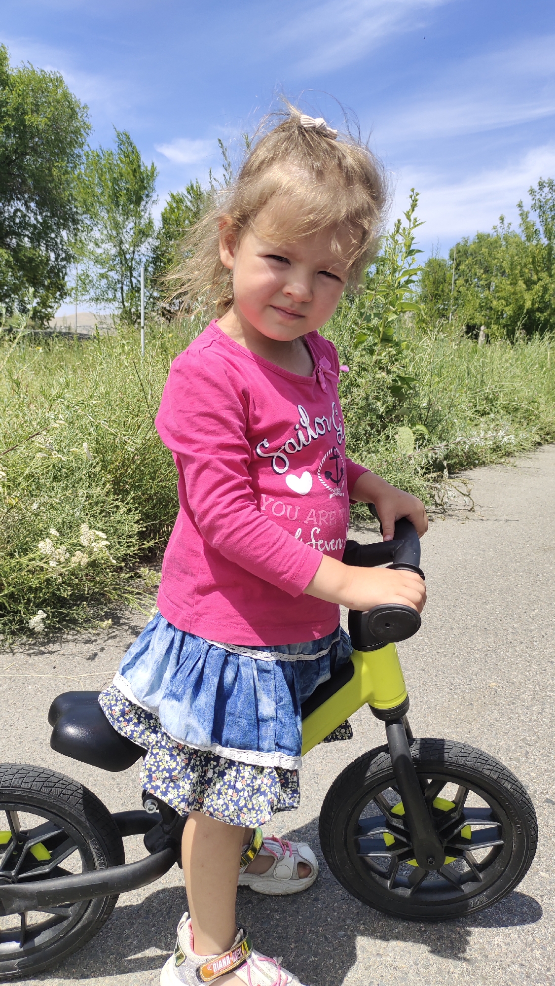 A young Caucasian girl with blonde hair pulled into a high ponytail with a light-colored hair tie is standing outdoors next to a yellow balance bike on a paved surface. The girl is wearing a bright pink long-sleeved t-shirt with a nautical design that includes the words "Sailor," a heart, an anchor, and "YOU ARE SO SEVEN". She is also wearing a blue denim skirt with ruffled tiers and a floral print underskirt. Her feet are adorned with white and pink sneakers with gold accents. She is holding the handlebars of the balance bike with her hands. The background consists of tall green grass and trees under a bright blue sky with scattered white clouds. The image was taken on a sunny day.