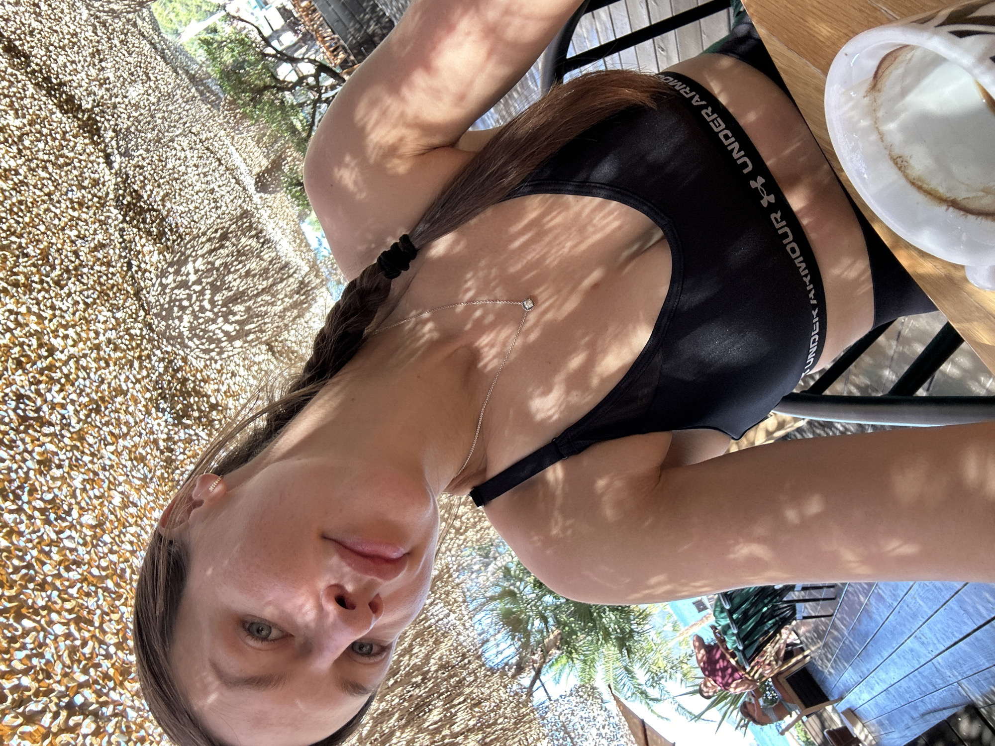 A woman with dark brown hair in a braid, wearing a black Under Armour sports bra and a silver necklace, takes a selfie outdoors. She has a faint smile and is looking towards the camera. The background shows a thatched awning casting dappled shadows, trees, and a body of water, suggesting a cafe or resort setting. A white cup with coffee foam is partially visible in the foreground. The woman appears to be seated at a wooden table. The location is described as Boreti, Montenegro, a coastal area known for its beaches and resorts.